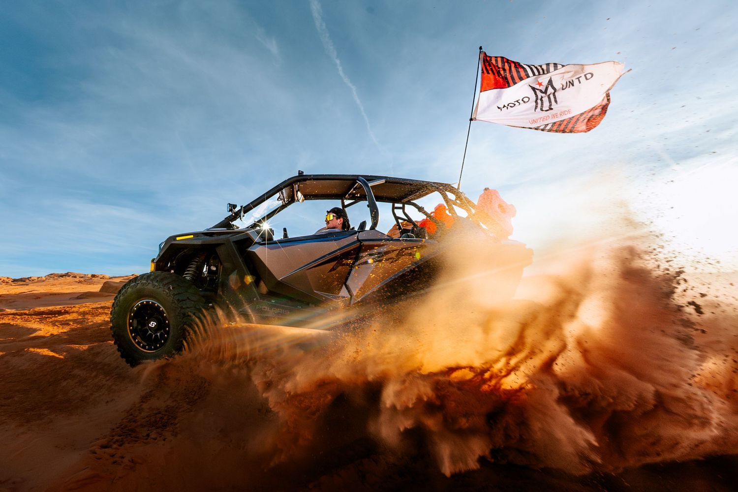 A UTV side-by-side kicks up red desert sand dunes while flying a branded flag under a blue sky.