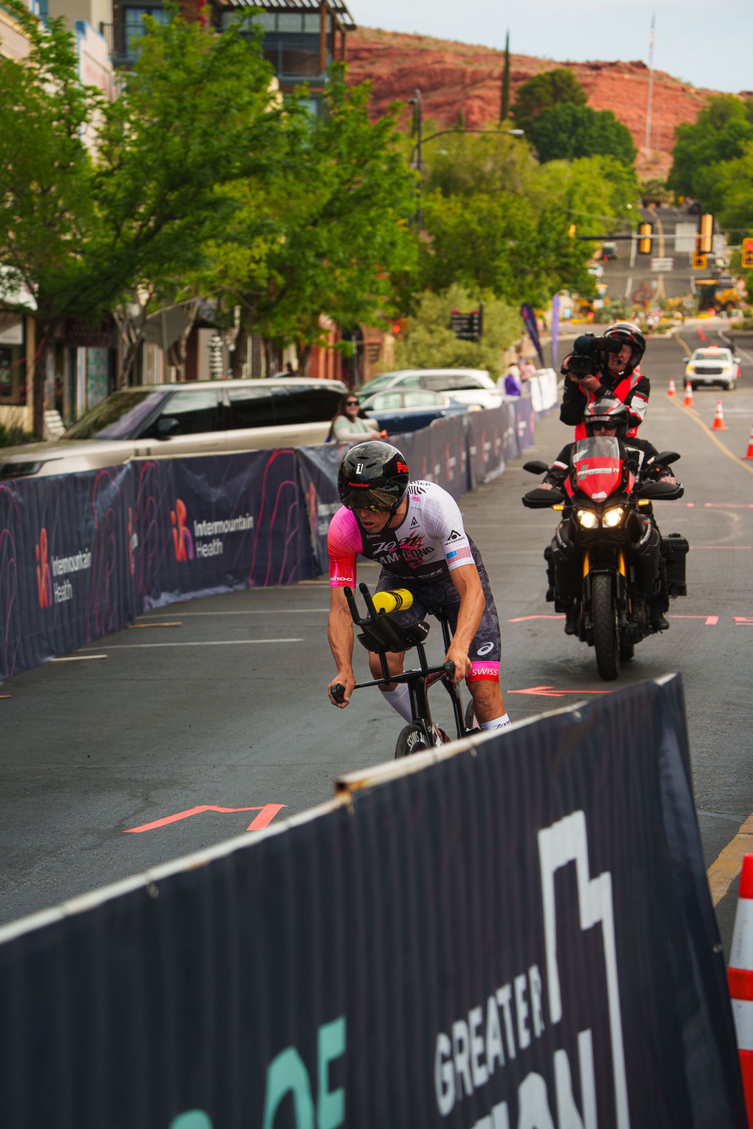 A triathlete in a pink helmet races on a time trial bike through city streets at an Ironman event.