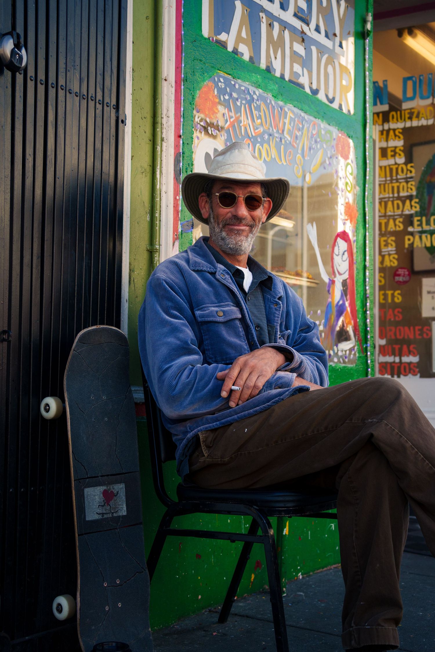 Smiling bearded man in a cowboy hat and sunglasses sitting outside a colorful Latin storefront with a skateboard.