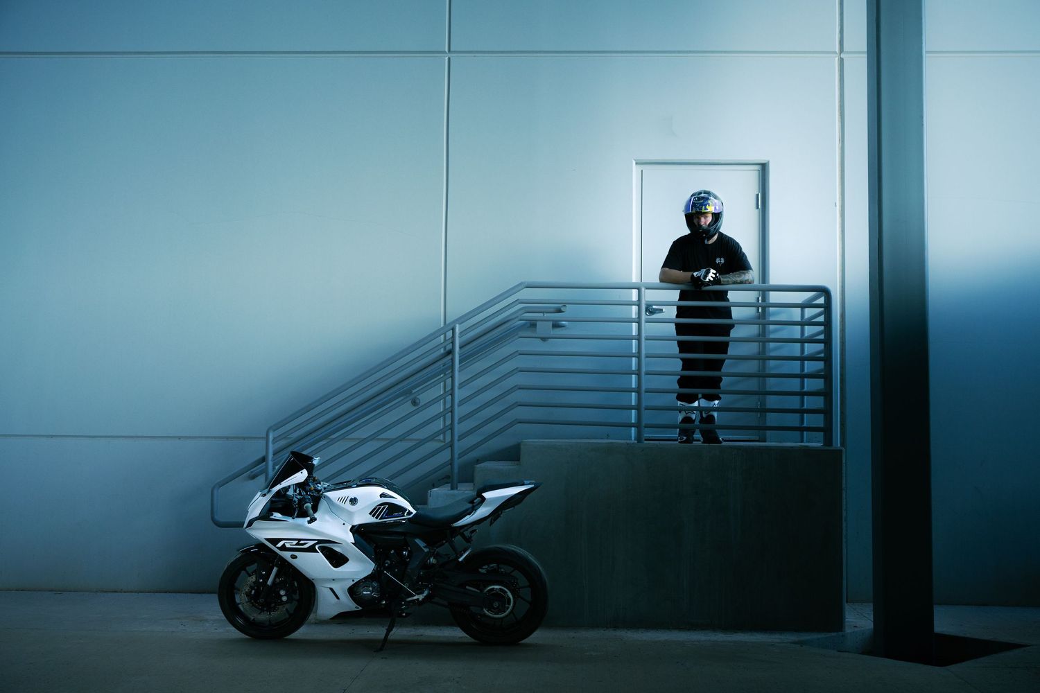 Helmeted rider standing on landing above white Yamaha R7 sport bike in dramatic blue-lit building.