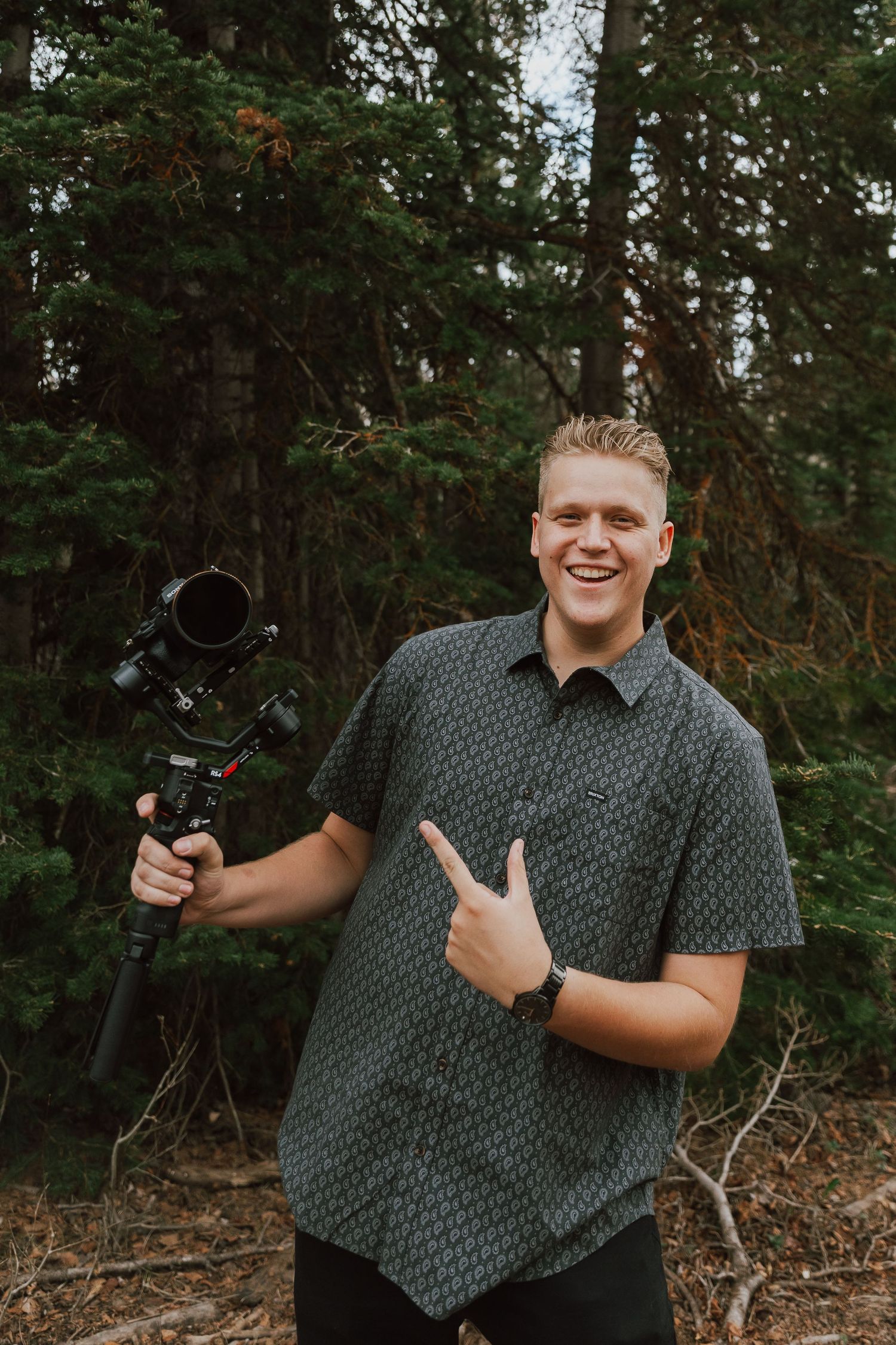 Smiling young videographer holds a professional camera gimbal stabilizer while posing in a pine forest.