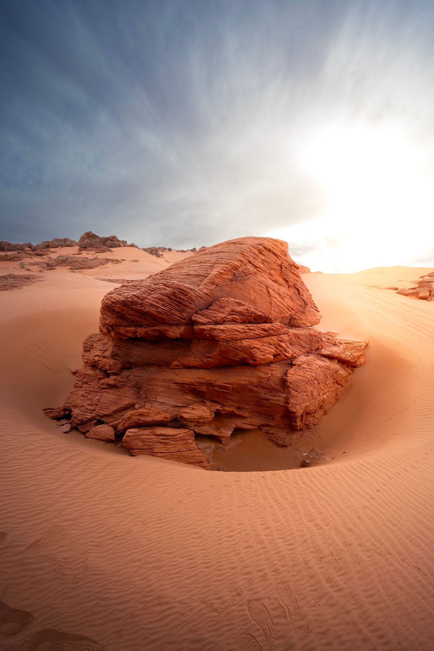 A large red sandstone rock formation rises from rippled sand dunes in a vast desert landscape under a dramatic sky.