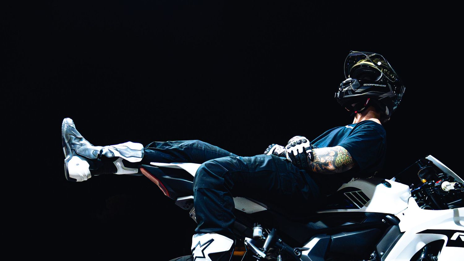 Tattooed motorcyclist in helmet reclines on white sport bike against dark background.