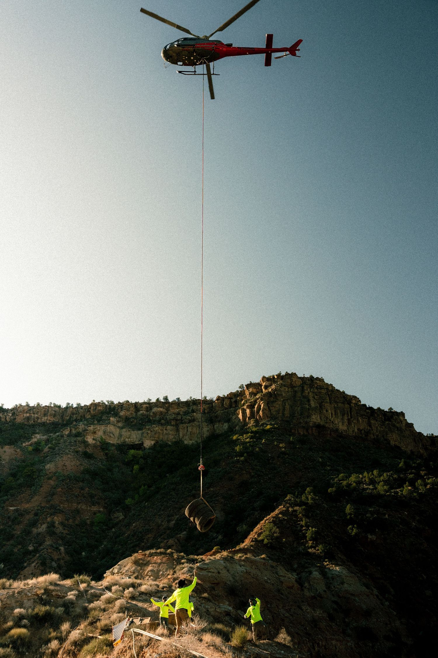 Red helicopter uses a long line to airlift supplies over a rugged canyon ridge with workers in neon vests below.