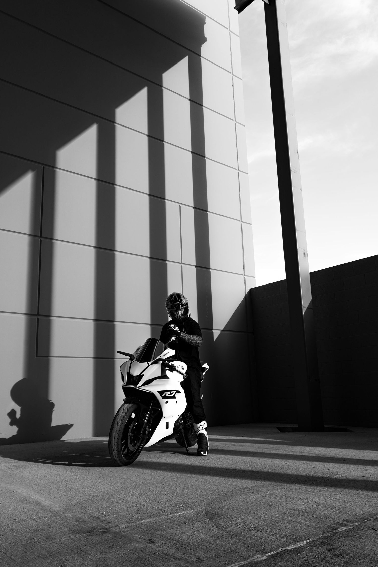 Black and white photo of helmeted motorcyclist on white Yamaha R7 beside modern building with dramatic shadows.