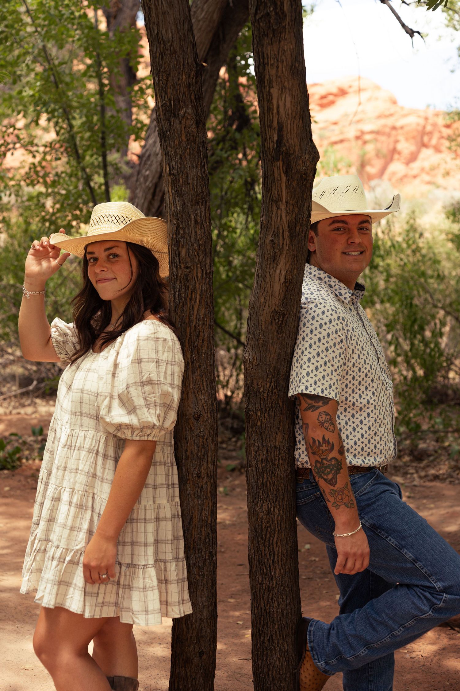 Couple in cowboy hats leaning against trees in a desert landscape with red rock formations behind them.