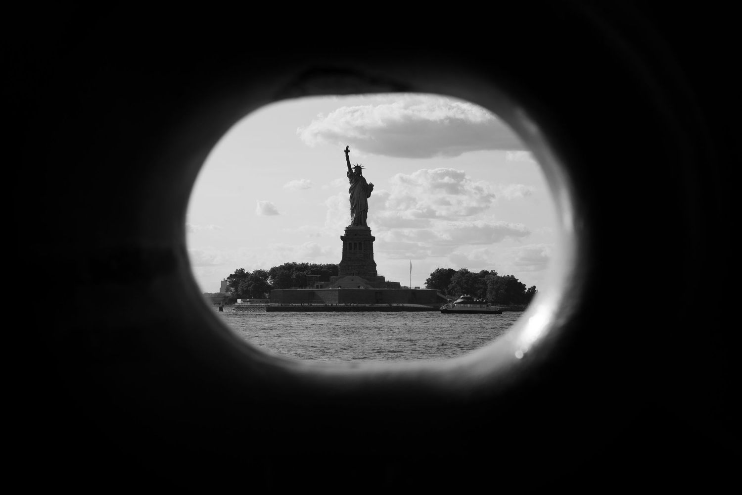 The Statue of Liberty framed through a circular porthole window in a dramatic black and white photograph.