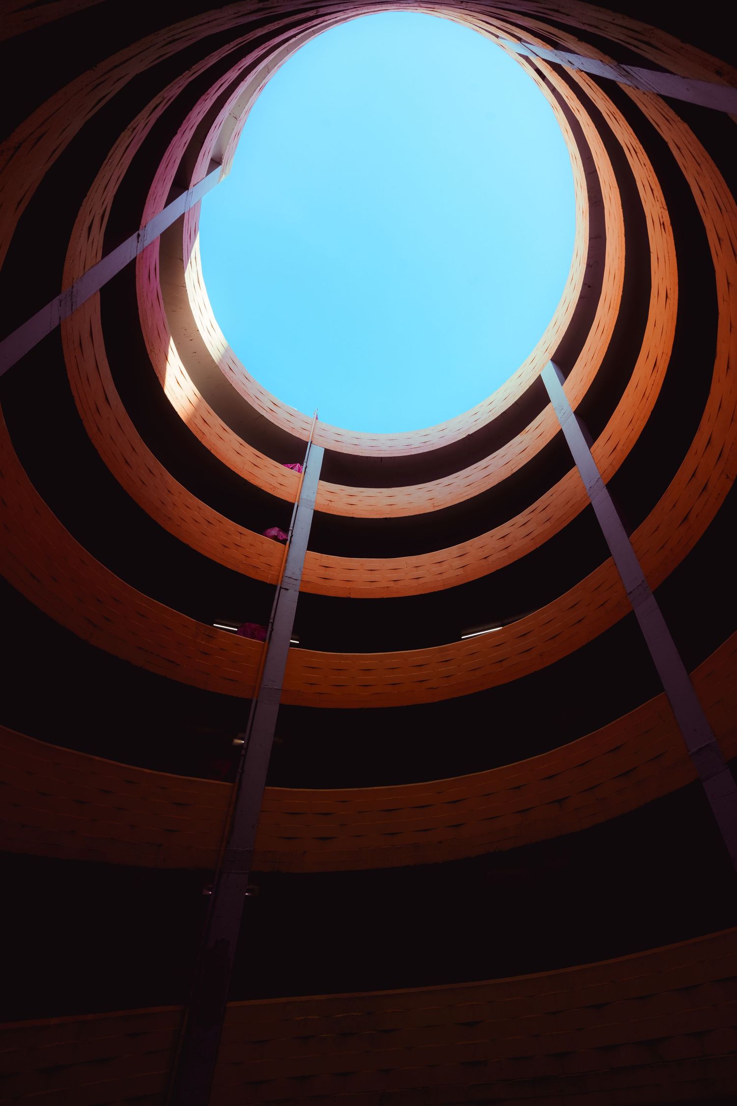 Low angle view looking up through a circular spiral wooden structure opening to a bright blue sky.
