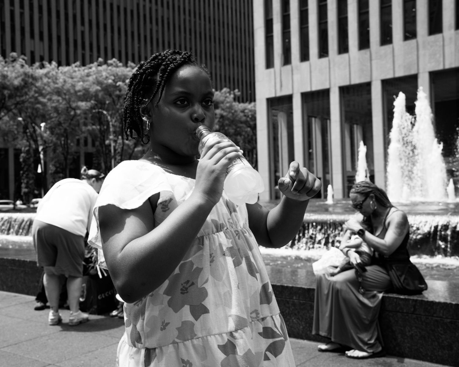 Black and white street photo of a young woman drinking from a cup near a city fountain plaza.
