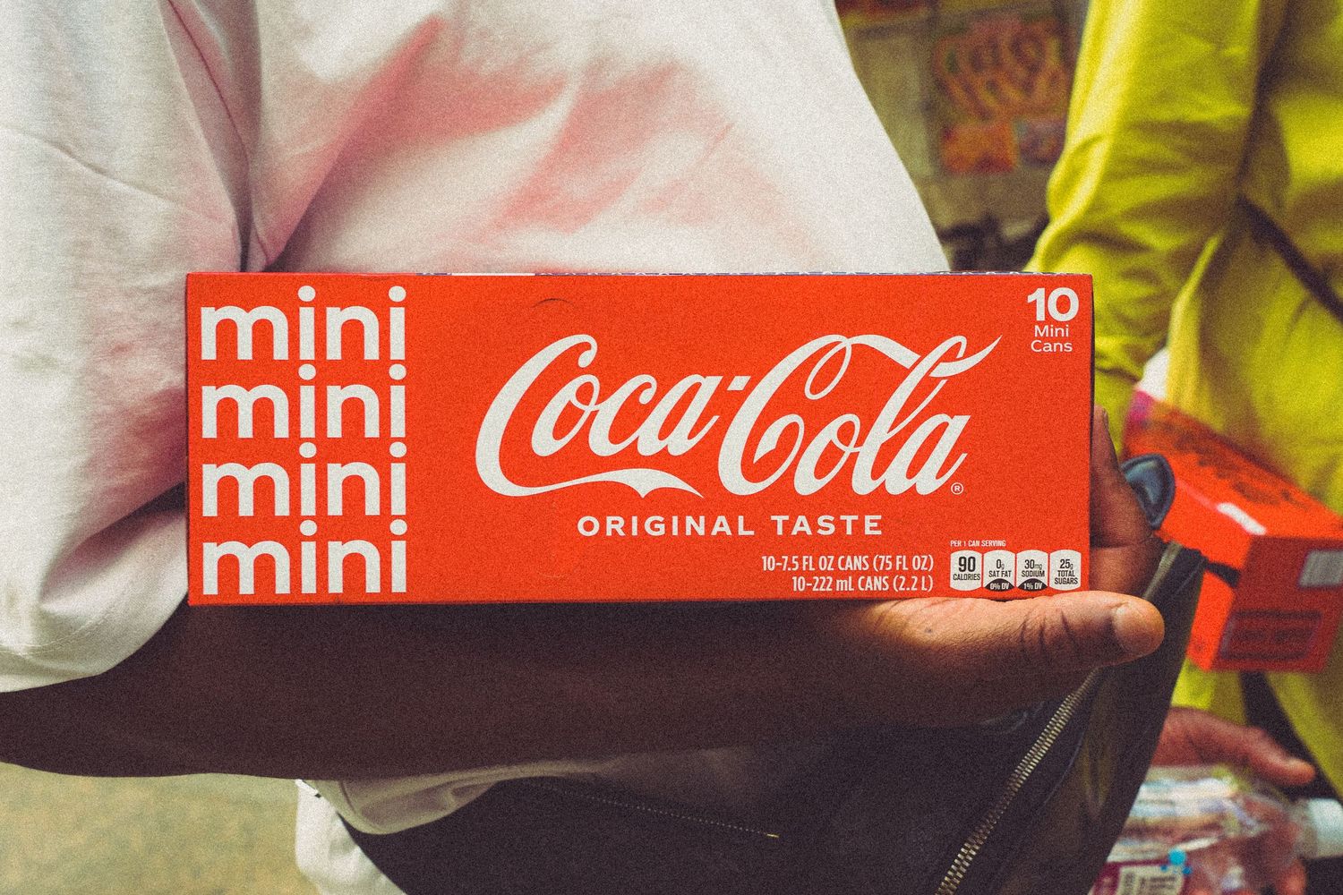 Person holding a box of Coca-Cola Mini cans with original taste branding in a store setting.