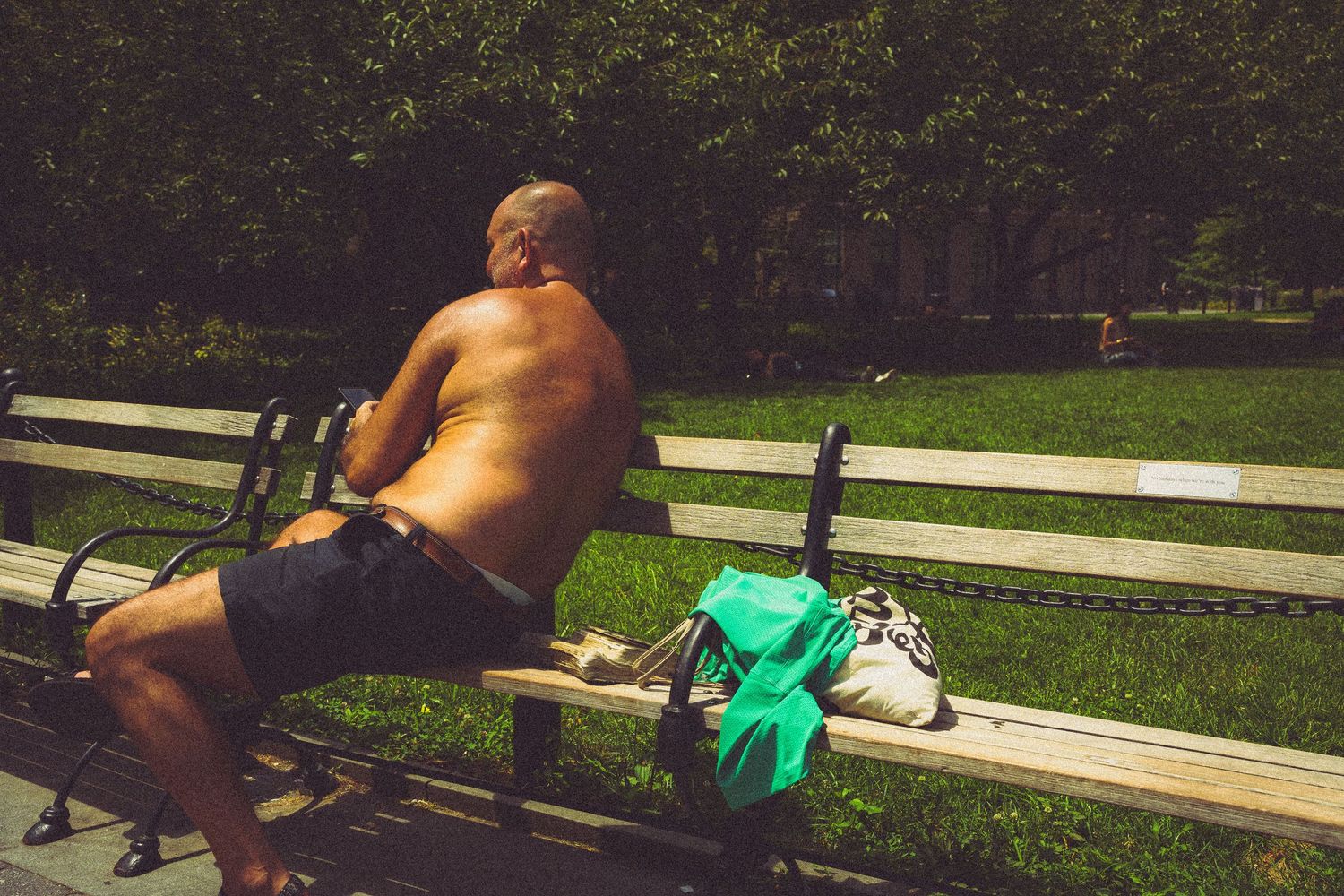 Shirtless man sits on park bench in sunny green park with backpack beside him.