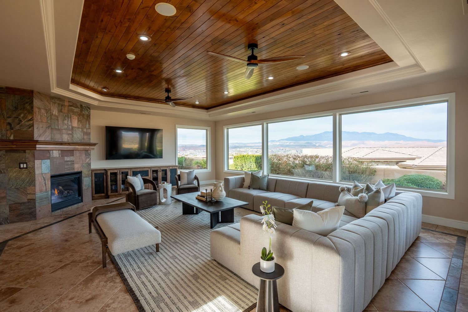 Luxury living room with wood tray ceiling, stone fireplace, large windows, mountain views, and neutral sectional sofa.