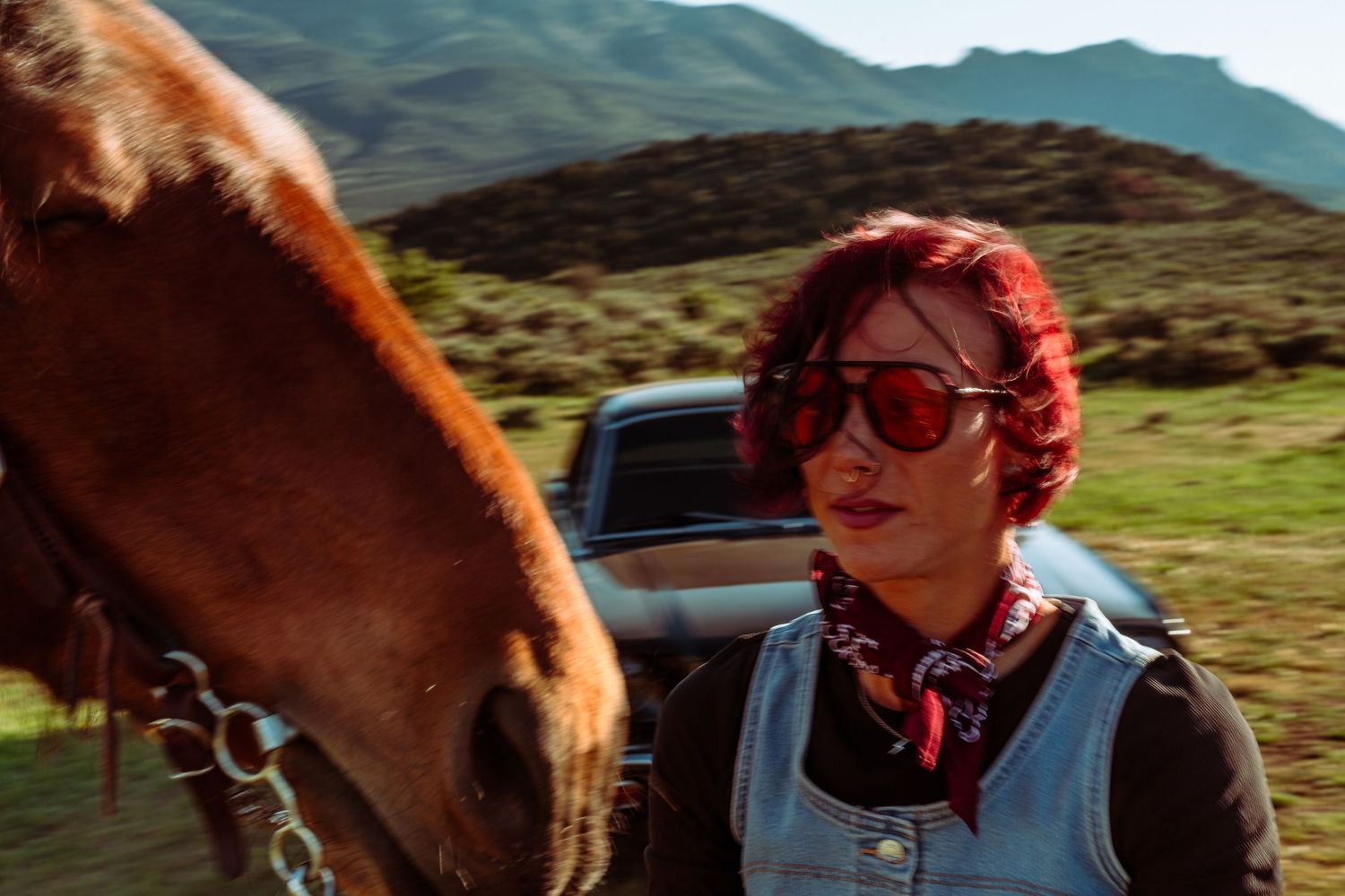 Woman with red hair and sunglasses standing beside a brown horse with mountains behind.