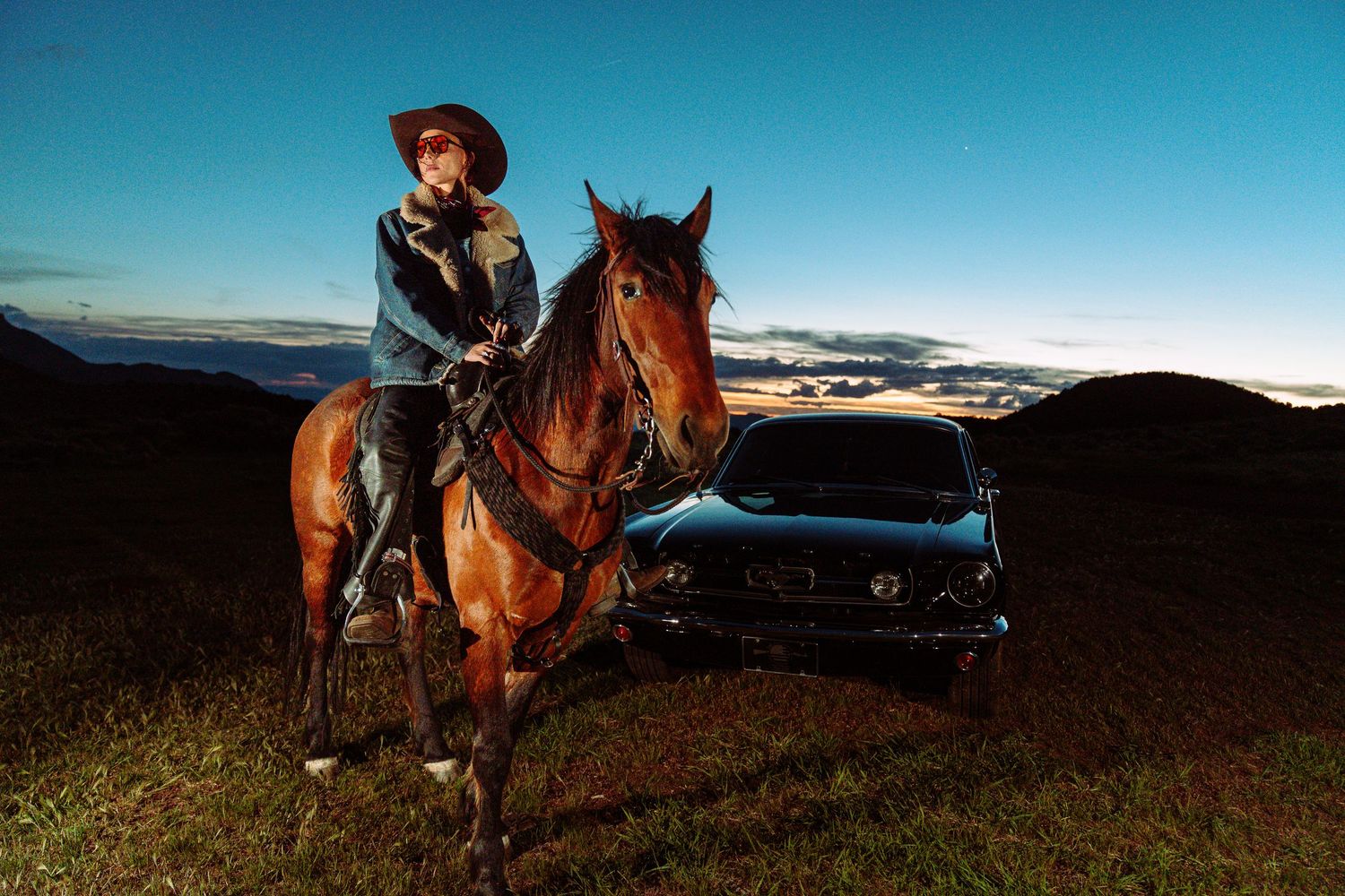 A cowboy in a hat rides a brown horse beside a classic black car on a scenic hillside at dusk.