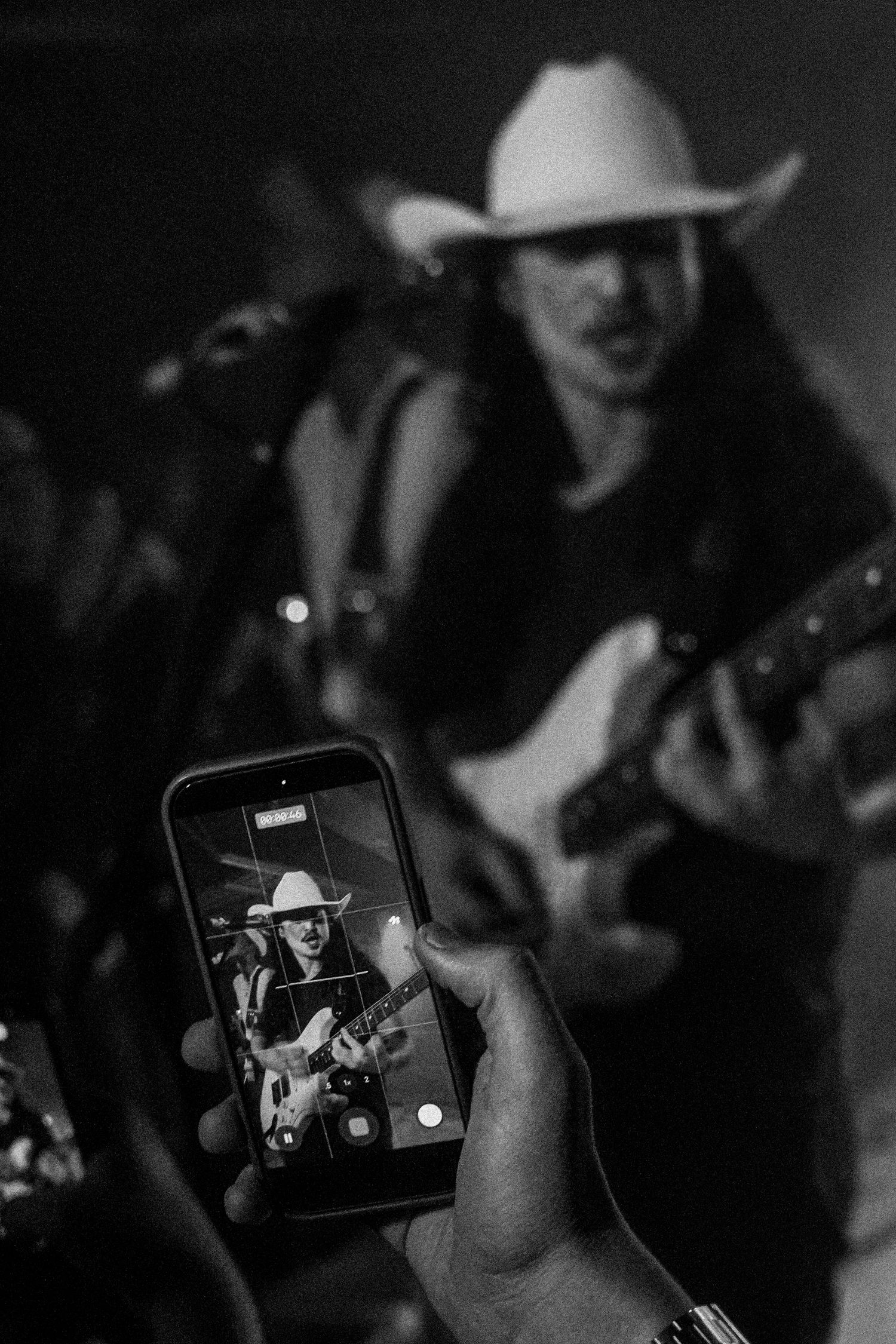 Audience member films a cowboy hat-wearing guitarist performing live on stage captured on a smartphone screen.