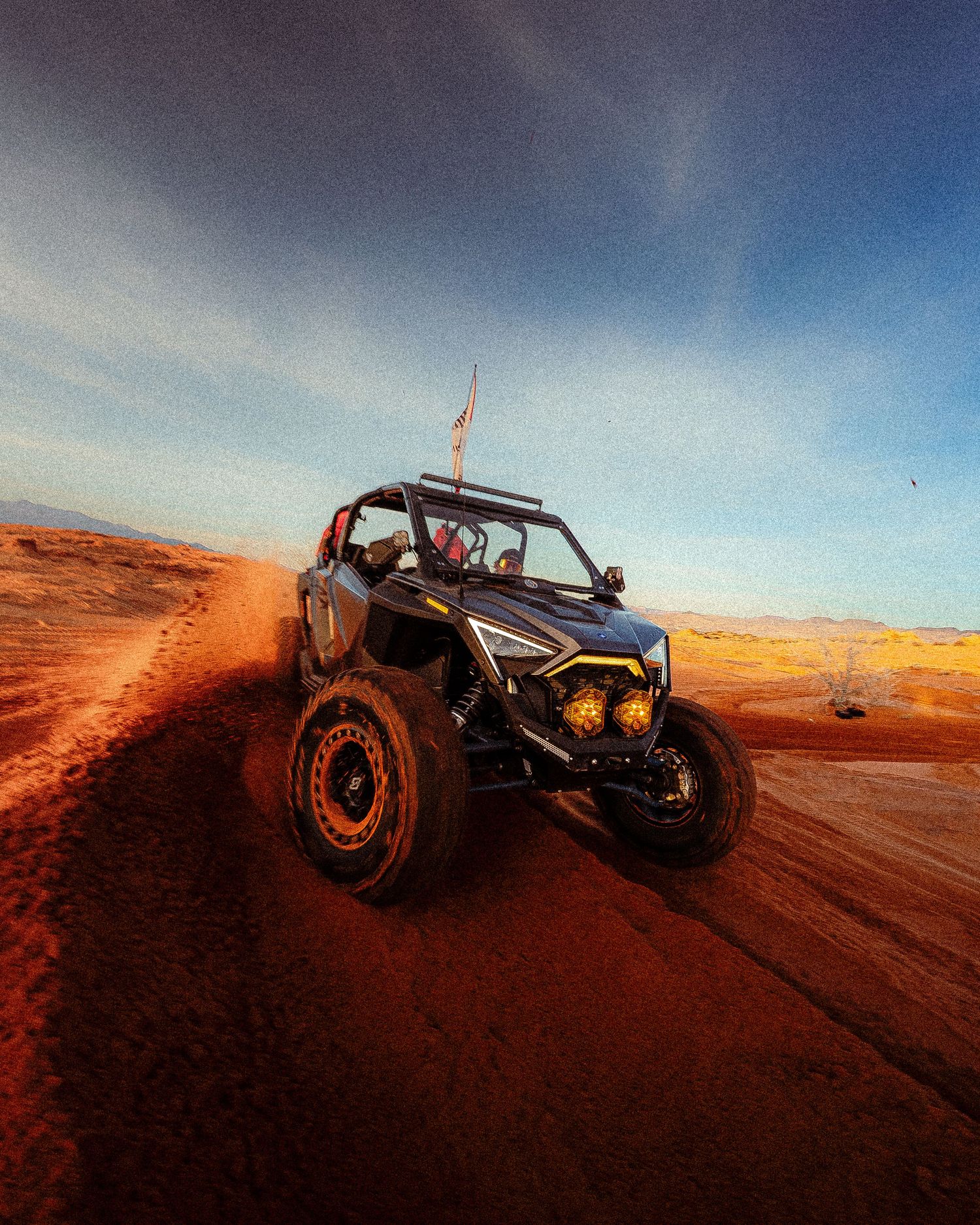 UTV side-by-side climbs red sand dunes at sunset with yellow auxiliary lights glowing brightly.
