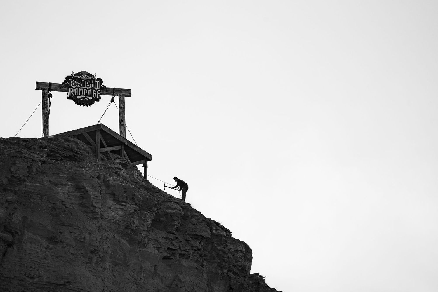 Silhouette of person climbing rocky cliff beside Red Bull Rampage sign in black and white photo.