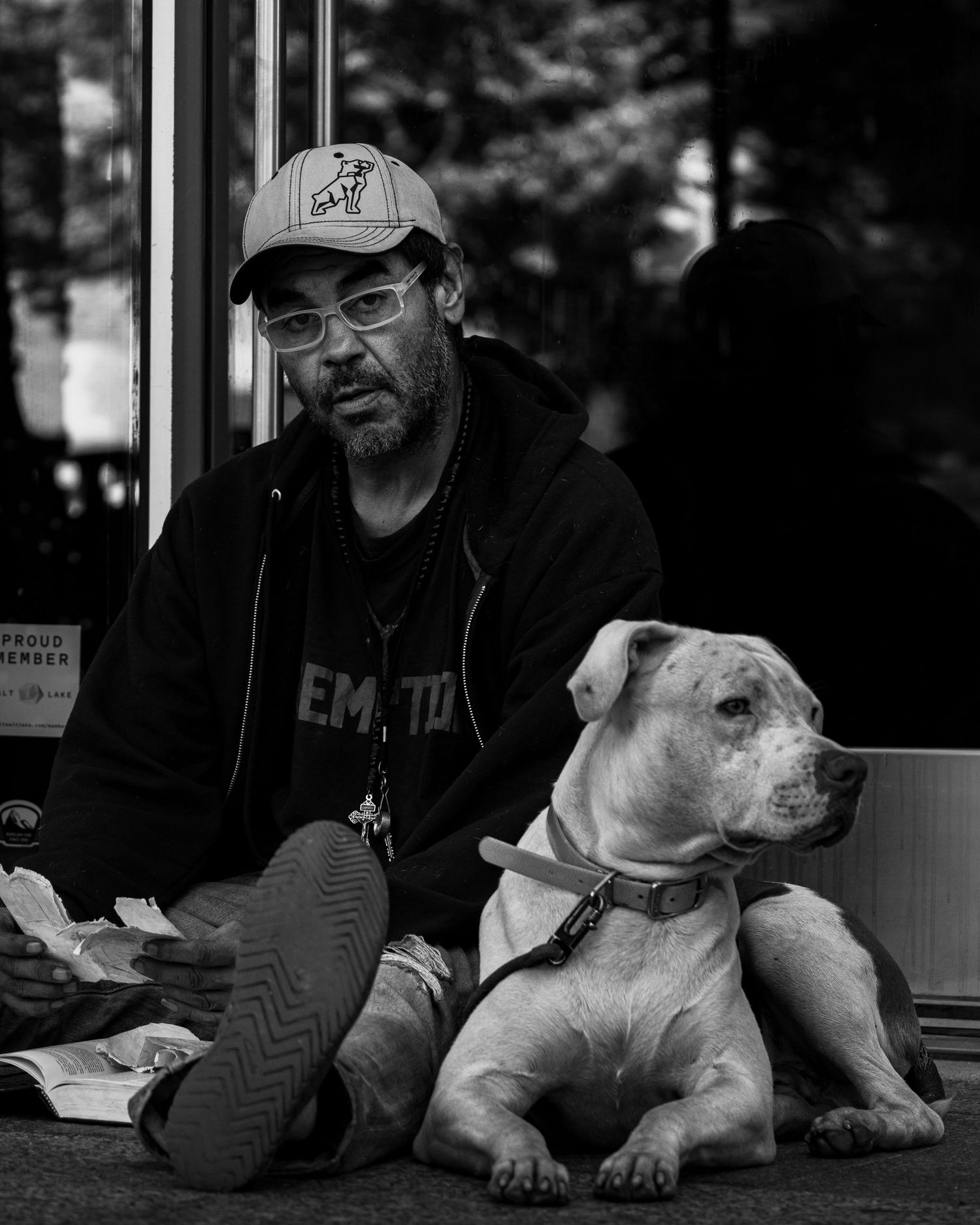 A bearded man wearing glasses and a cap sits beside a white pit bull dog on a sidewalk in black and white.