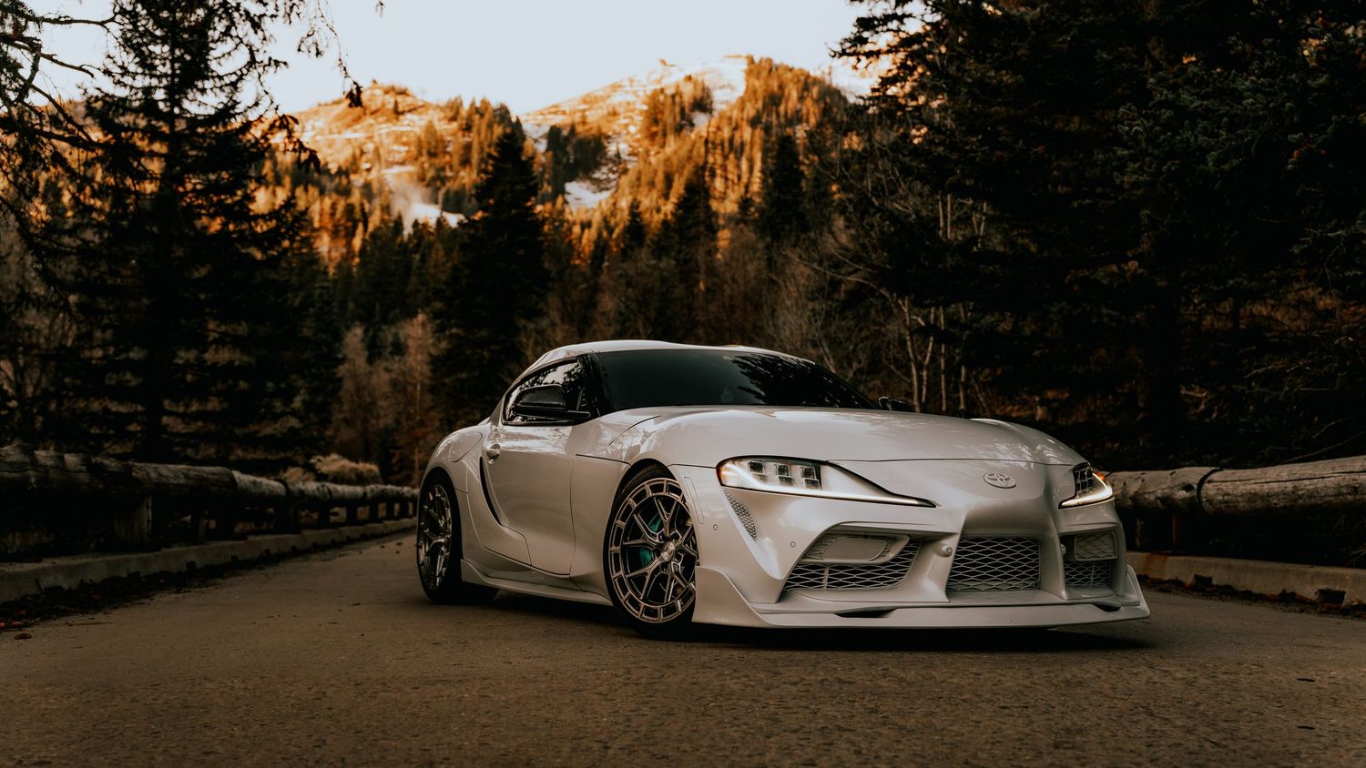 White Toyota Supra with wide body kit parked on mountain road surrounded by autumn trees at golden hour.