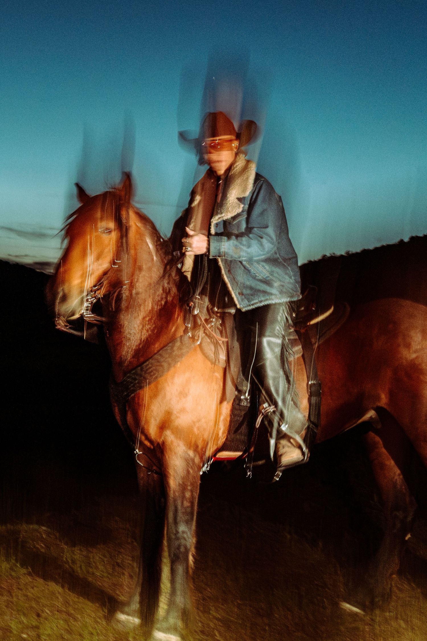 A cowboy in a denim jacket and hat rides a chestnut horse at dusk, captured with a motion blur effect.