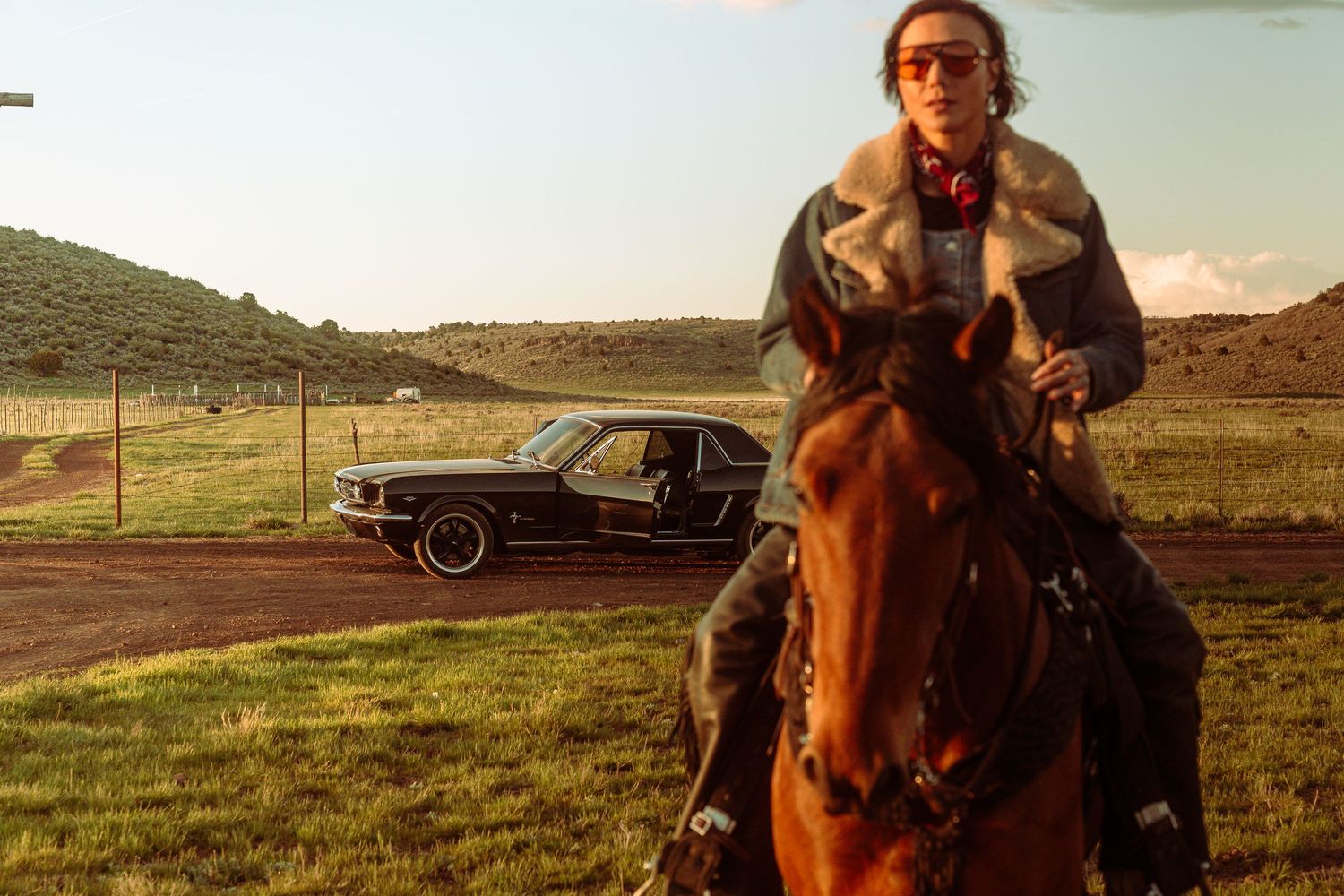 Woman on horseback rides past a classic black Ford Mustang on a rural dirt track at sunset.