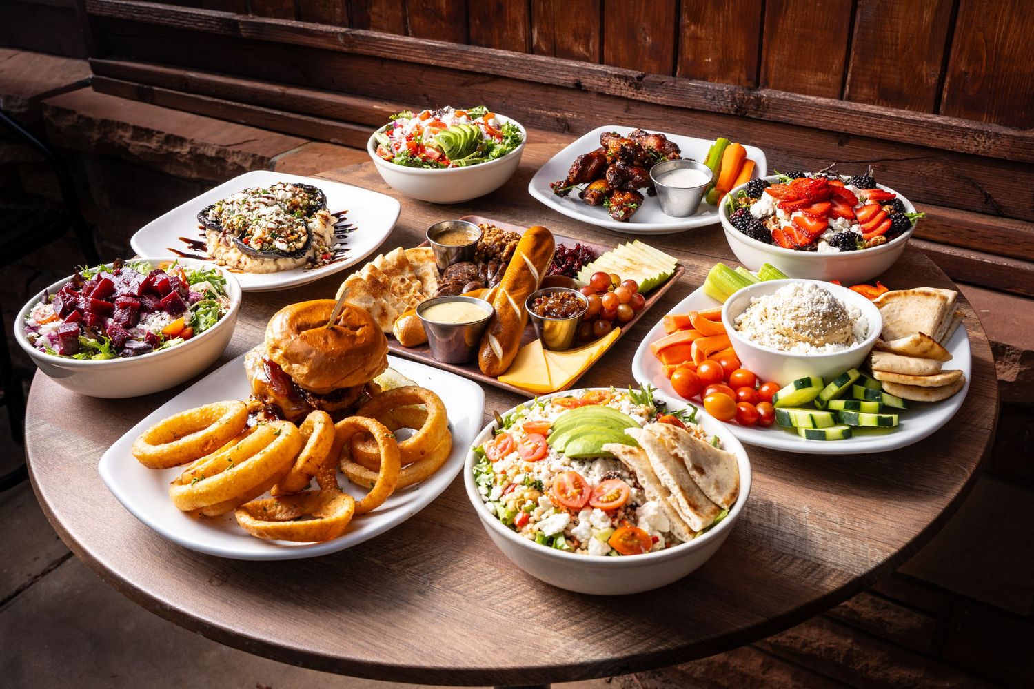 Overhead spread of diverse dishes including onion rings, salads, grilled meats, and hummus on round table.