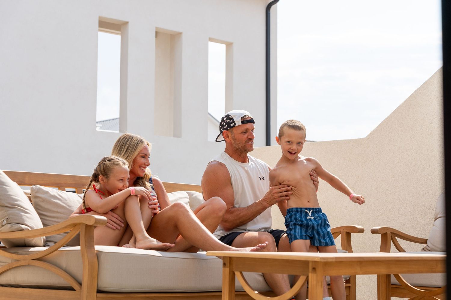 Happy family relaxing on an outdoor patio sofa at a modern resort, with a child jumping playfully.