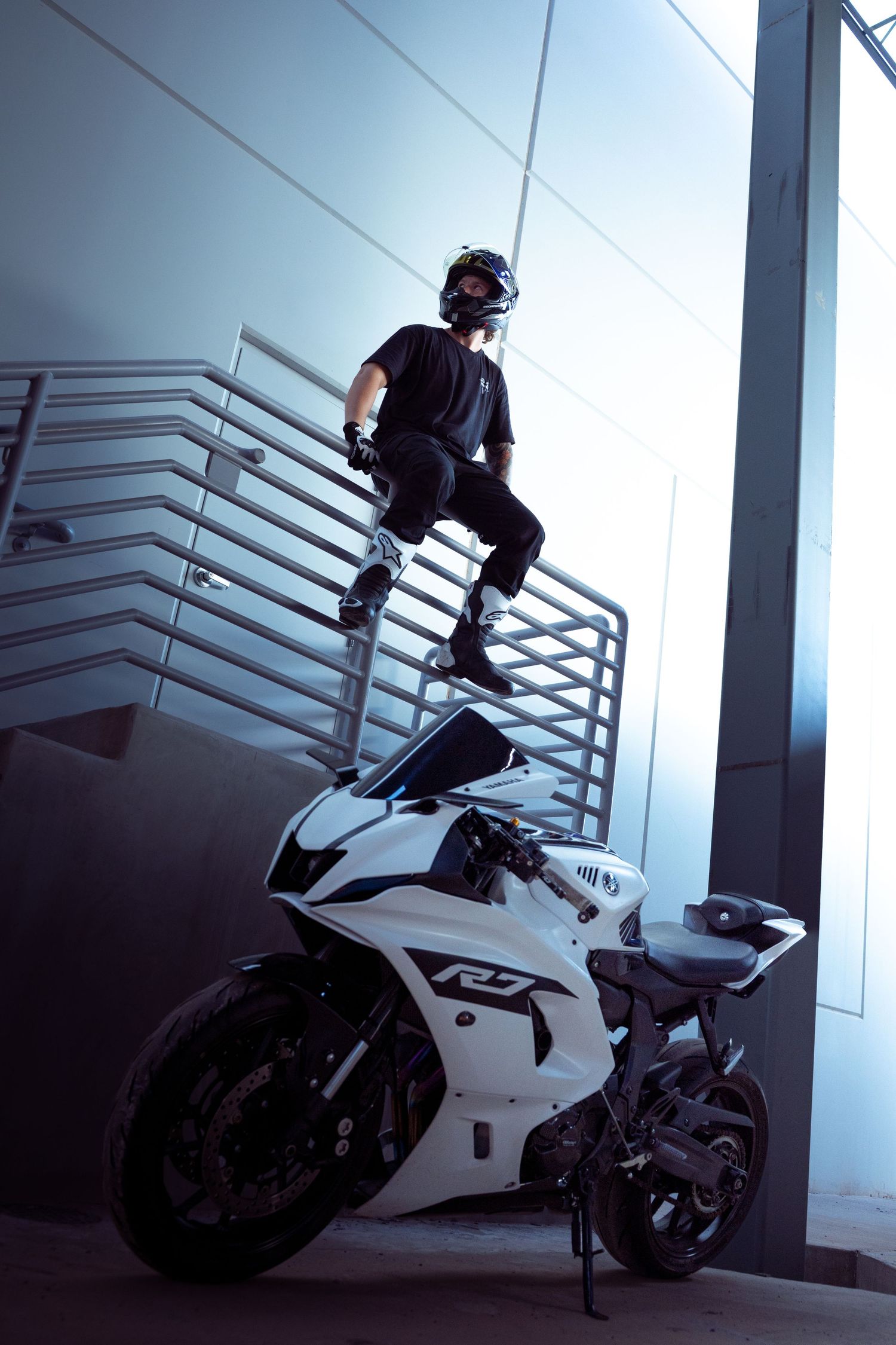 Motorcyclist in black helmet standing on white Yamaha R7 sport bike near indoor metal staircase.
