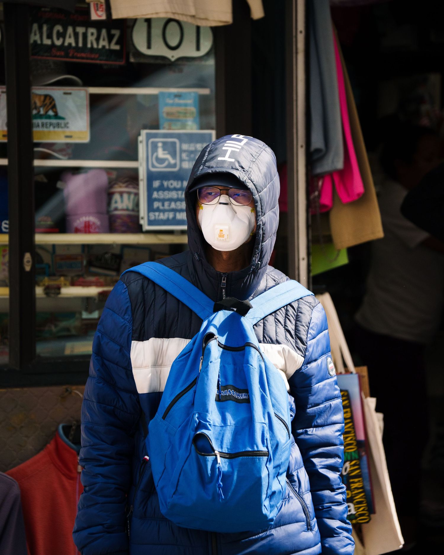 Person wearing N95 mask, blue jacket, and backpack standing outside Alcatraz souvenir shop in San Francisco.
