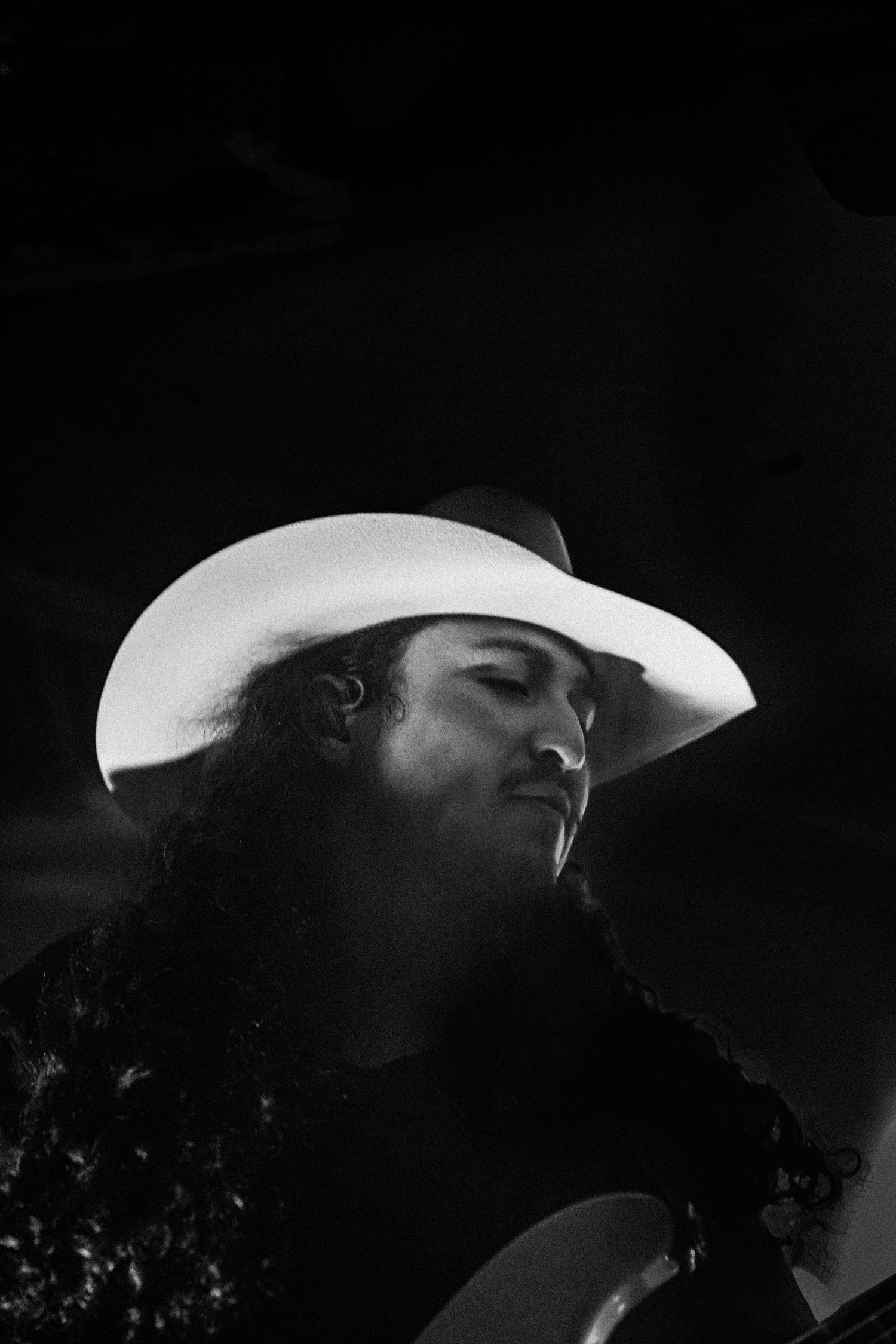 Dramatic black and white close-up portrait of a curly-haired musician wearing a glowing white cowboy hat.