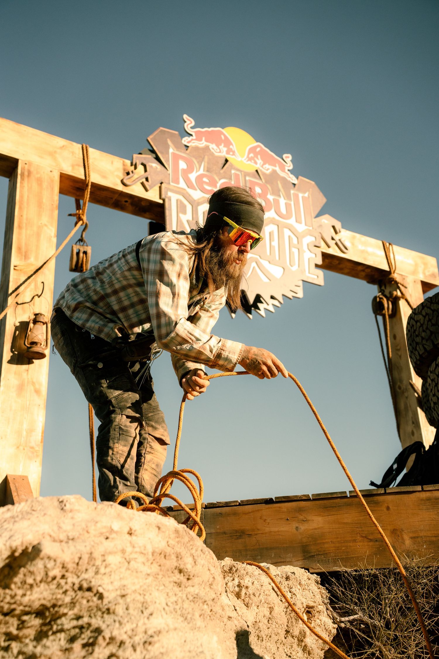 Bearded man with goggles and turban handles rope near Red Bull Rampage wooden sign on rocky terrain.