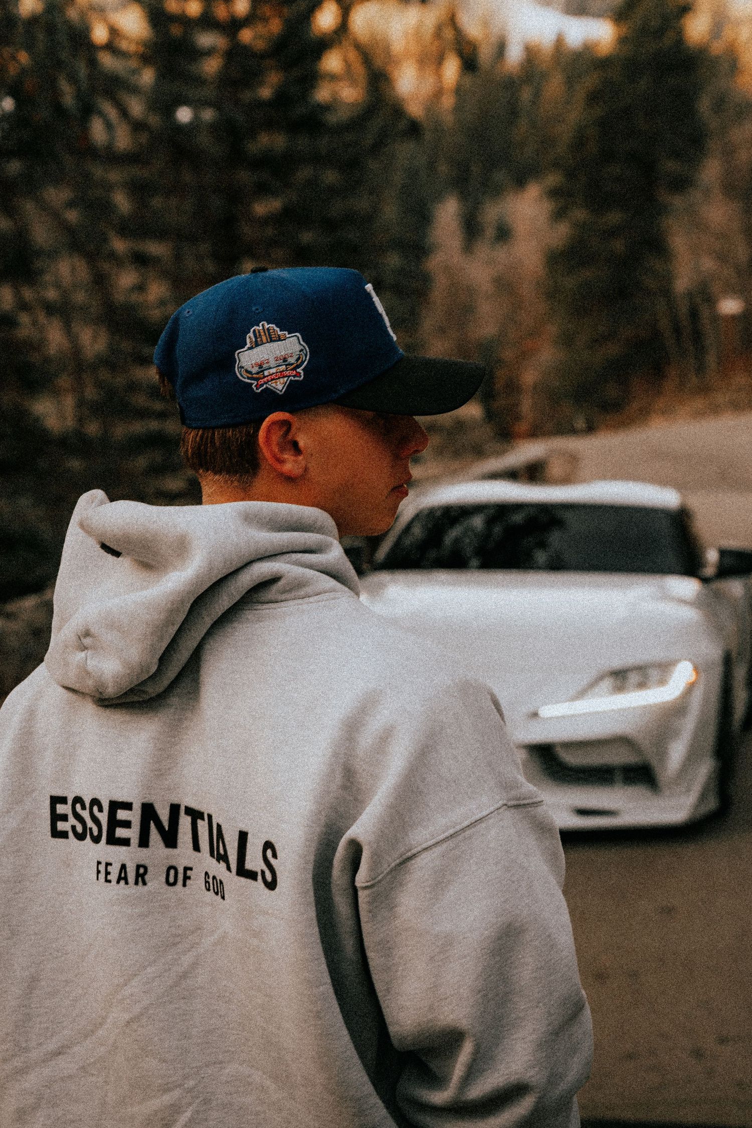 Man in gray Essentials Fear of God hoodie and blue snapback cap standing before a white sports car outdoors.
