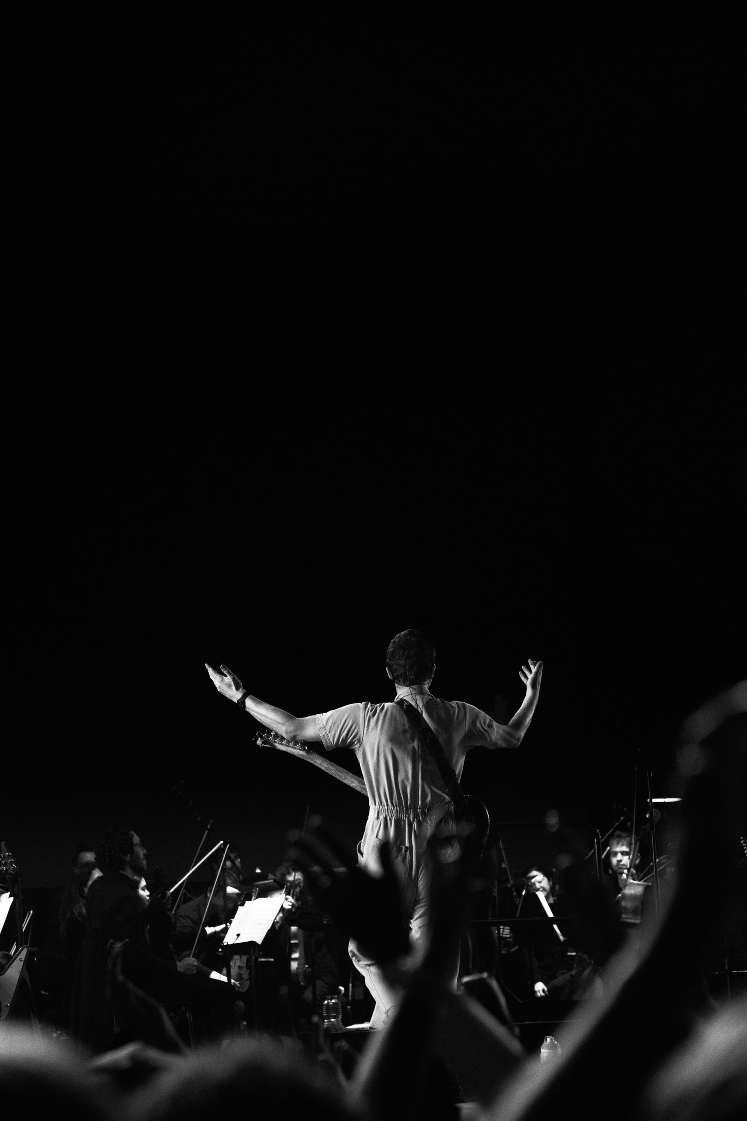 Black and white silhouette of a conductor with arms raised before an orchestra and crowd at night.
