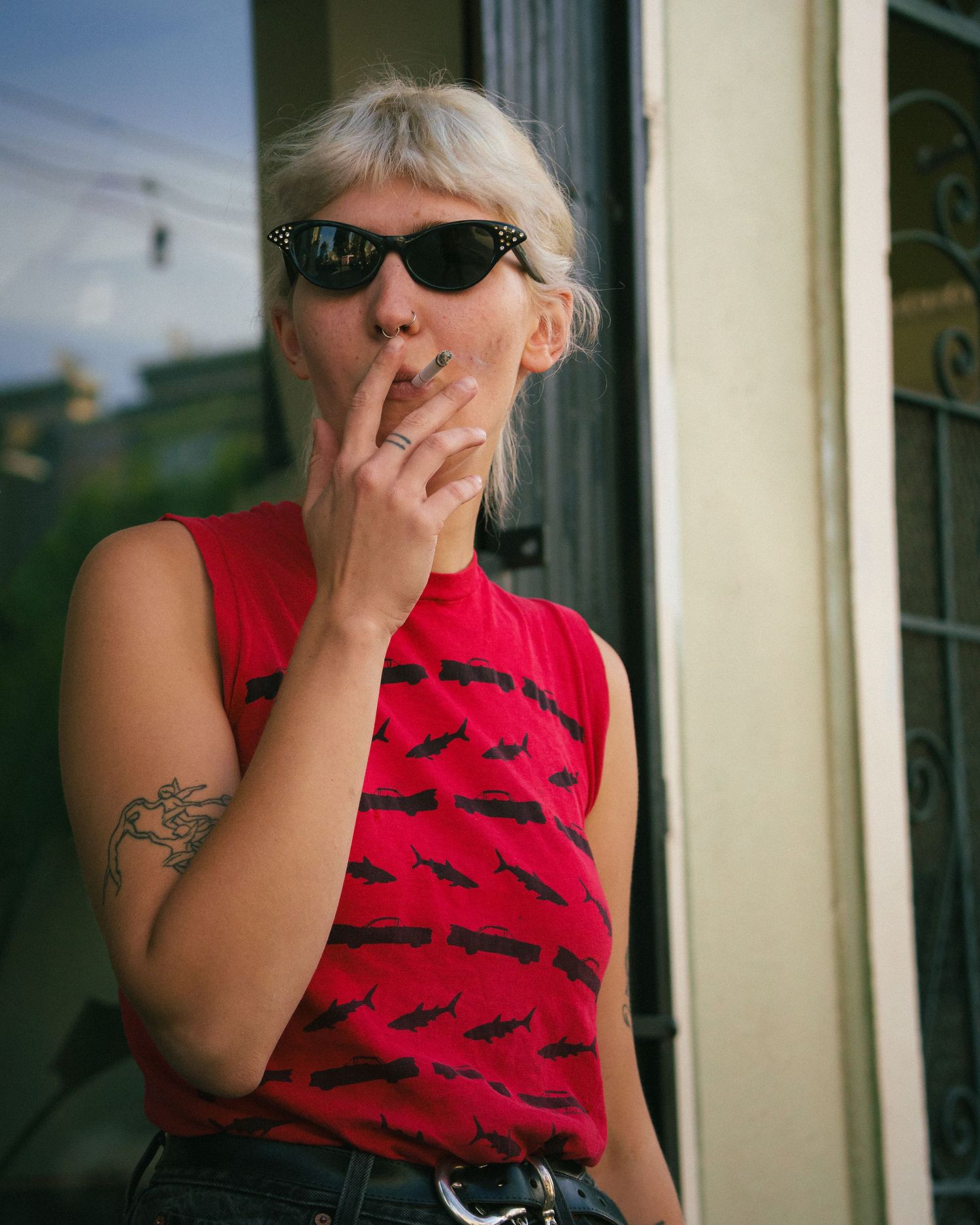 Woman in red sleeveless top and black sunglasses smoking a cigarette outdoors.