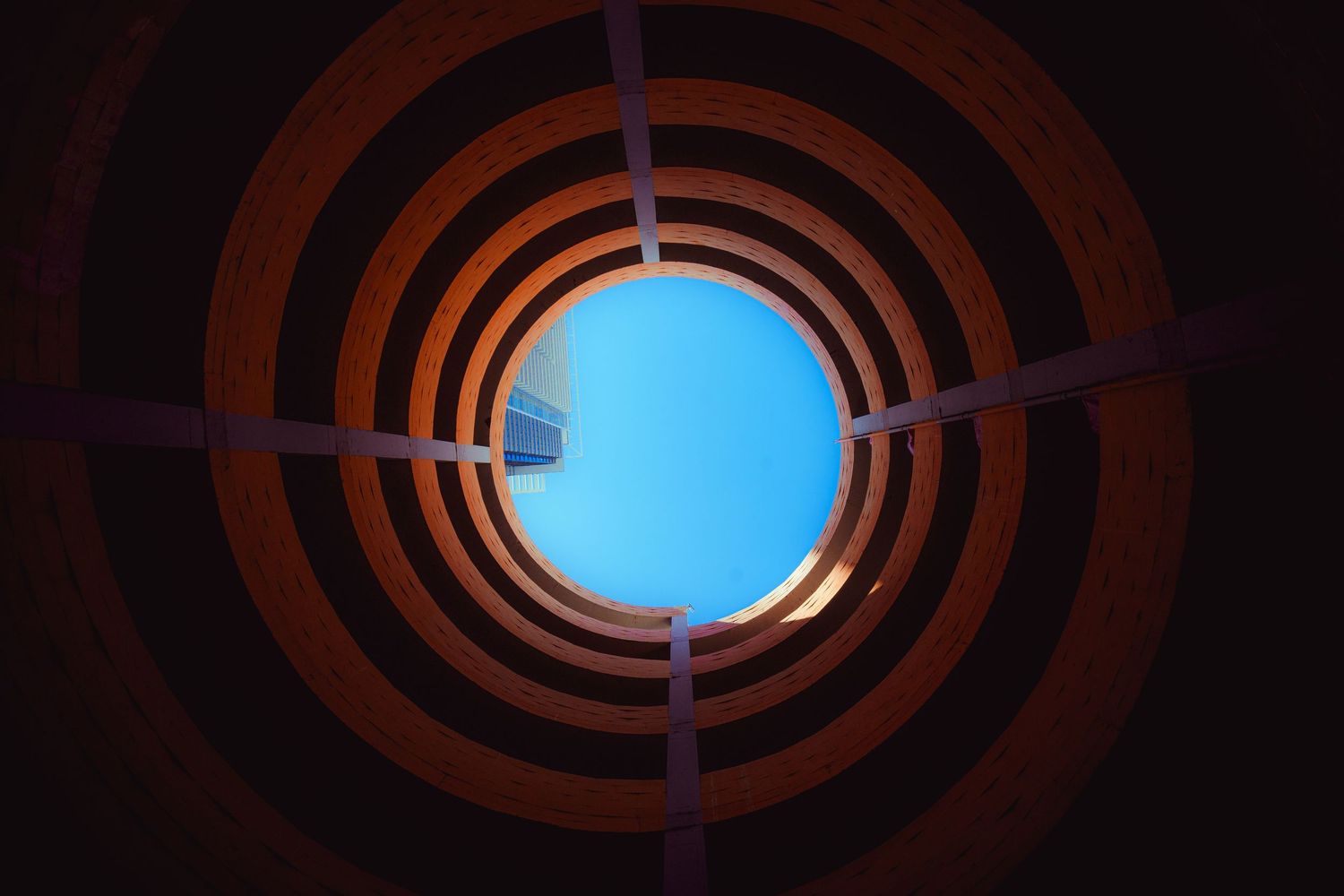 Looking up through a circular spiral architecture opening to a blue sky.