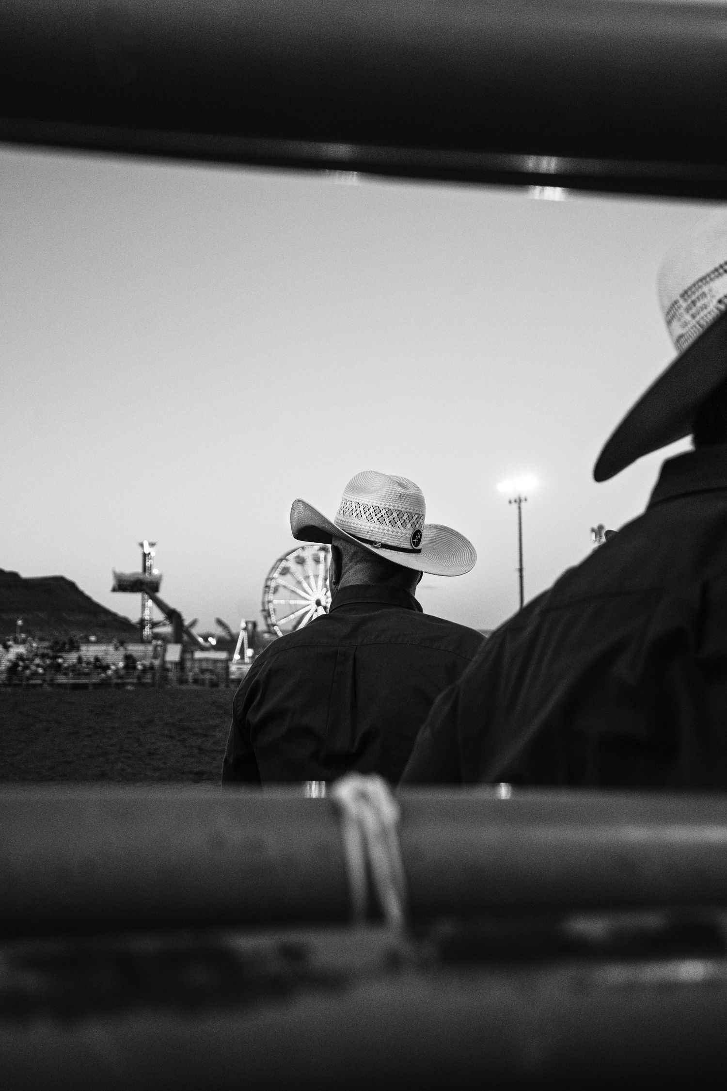 Black and white silhouette of cowboys watching a rodeo at dusk framed through metal chute rails.