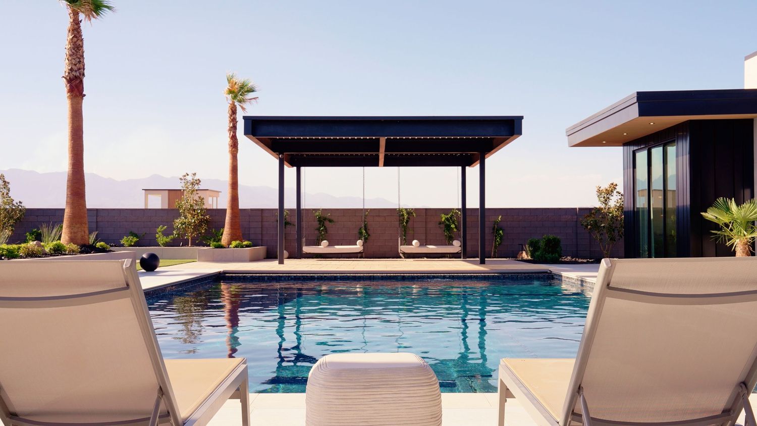 Modern luxury pool with lounge chairs, pergola, and palm trees in a desert setting.