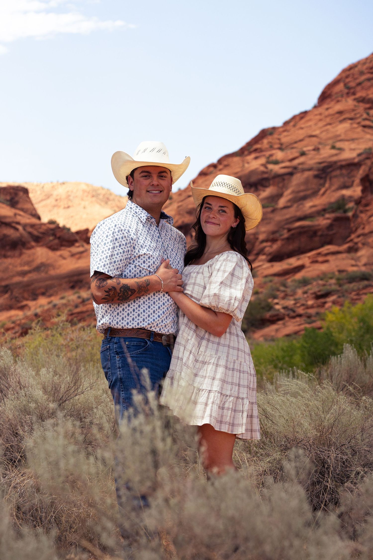 Couple in straw cowboy hats embracing in desert sagebrush with red rock cliffs.