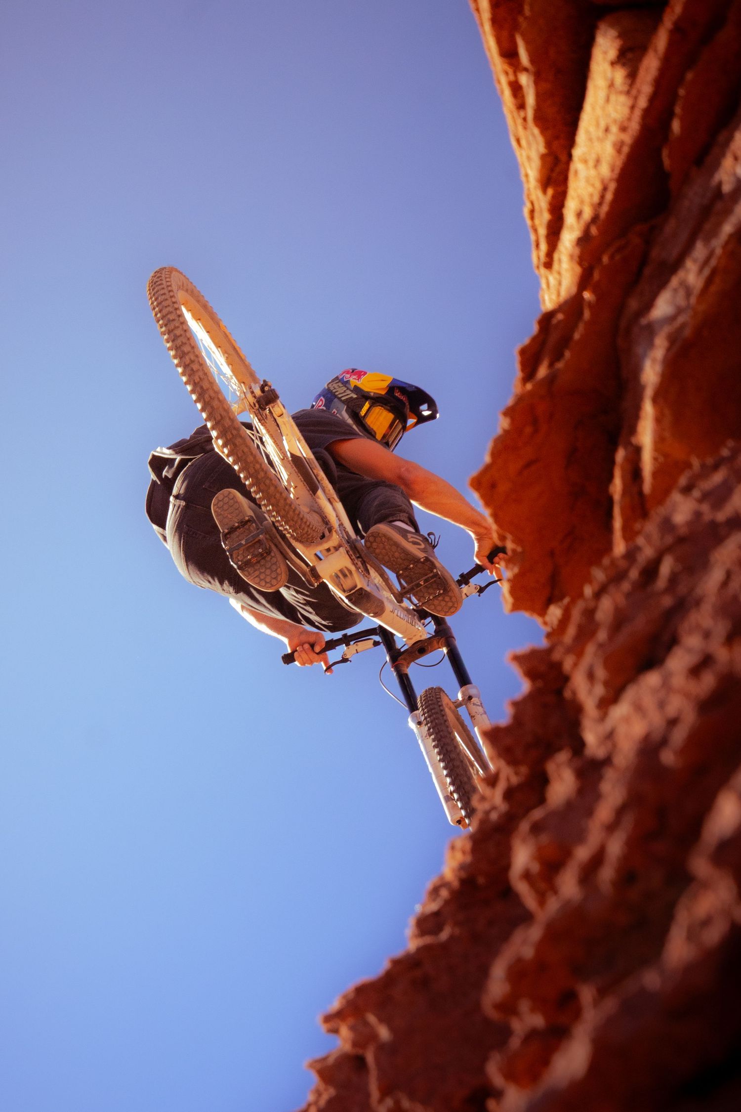 Mountain biker performing a daring drop off a red rock cliff face against a clear blue sky.