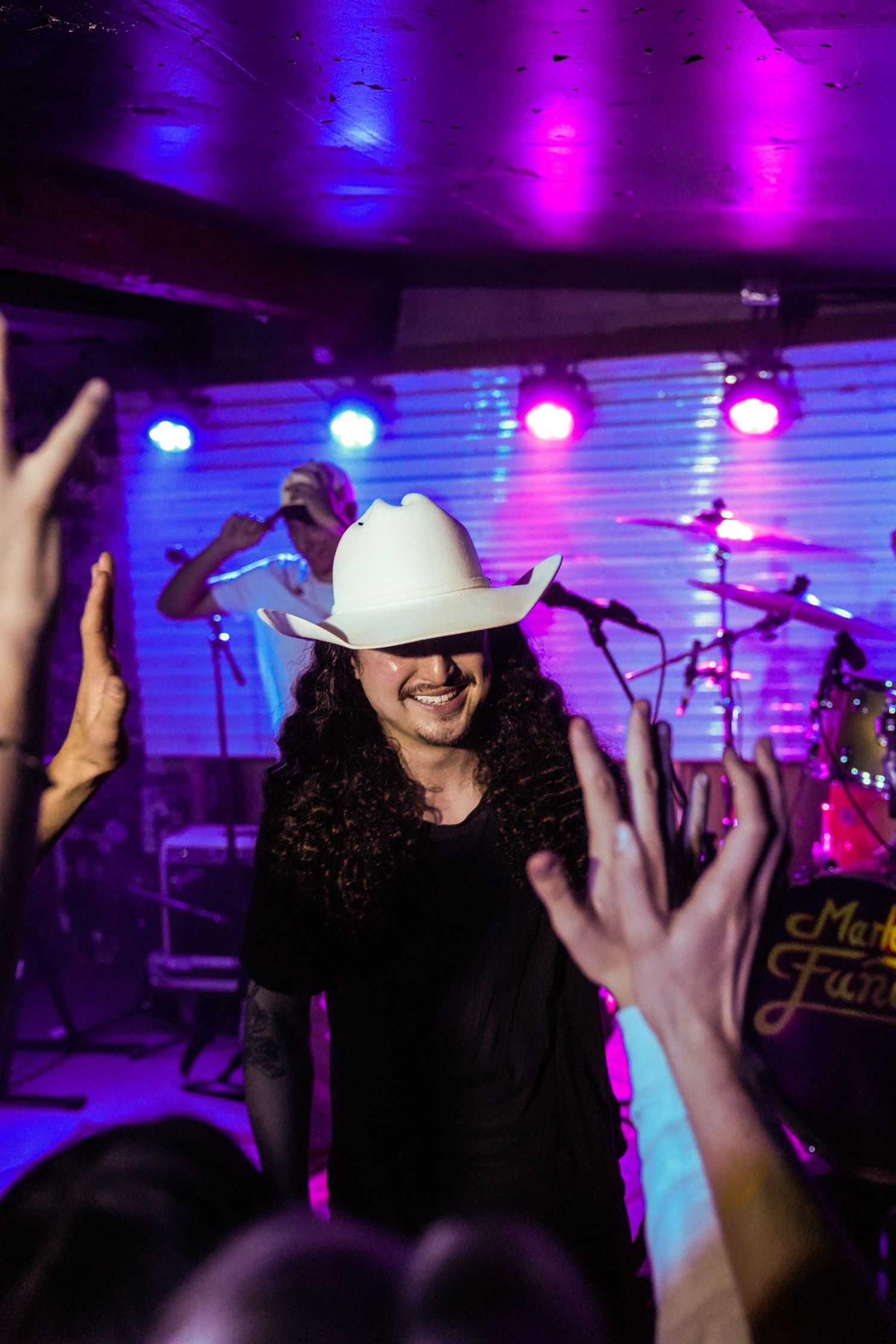 Energetic performer in a white cowboy hat smiles as excited fans reach toward the colorfully lit stage.