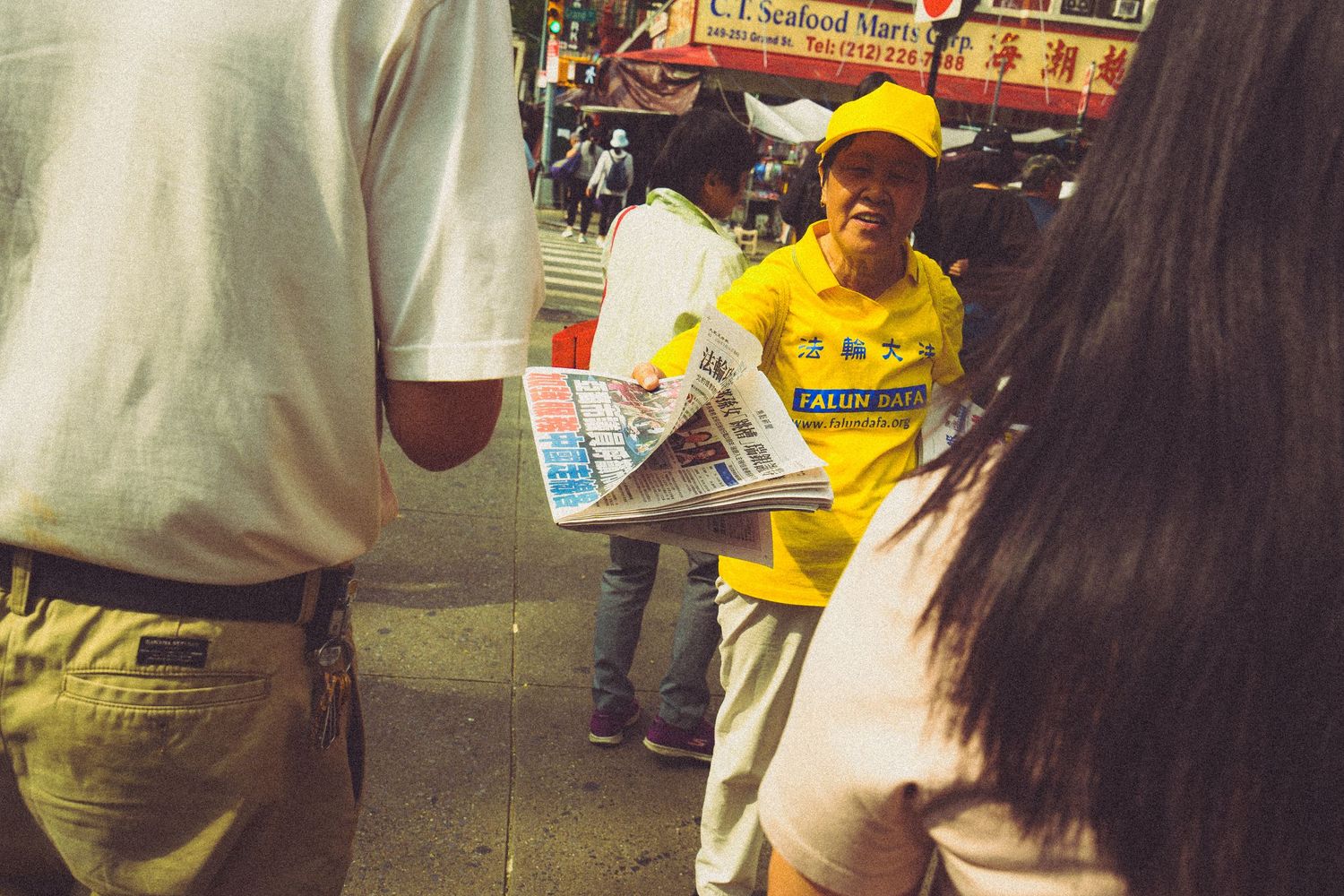 Falun Dafa member in yellow shirt distributes newspapers on a crowded Chinatown sidewalk.
