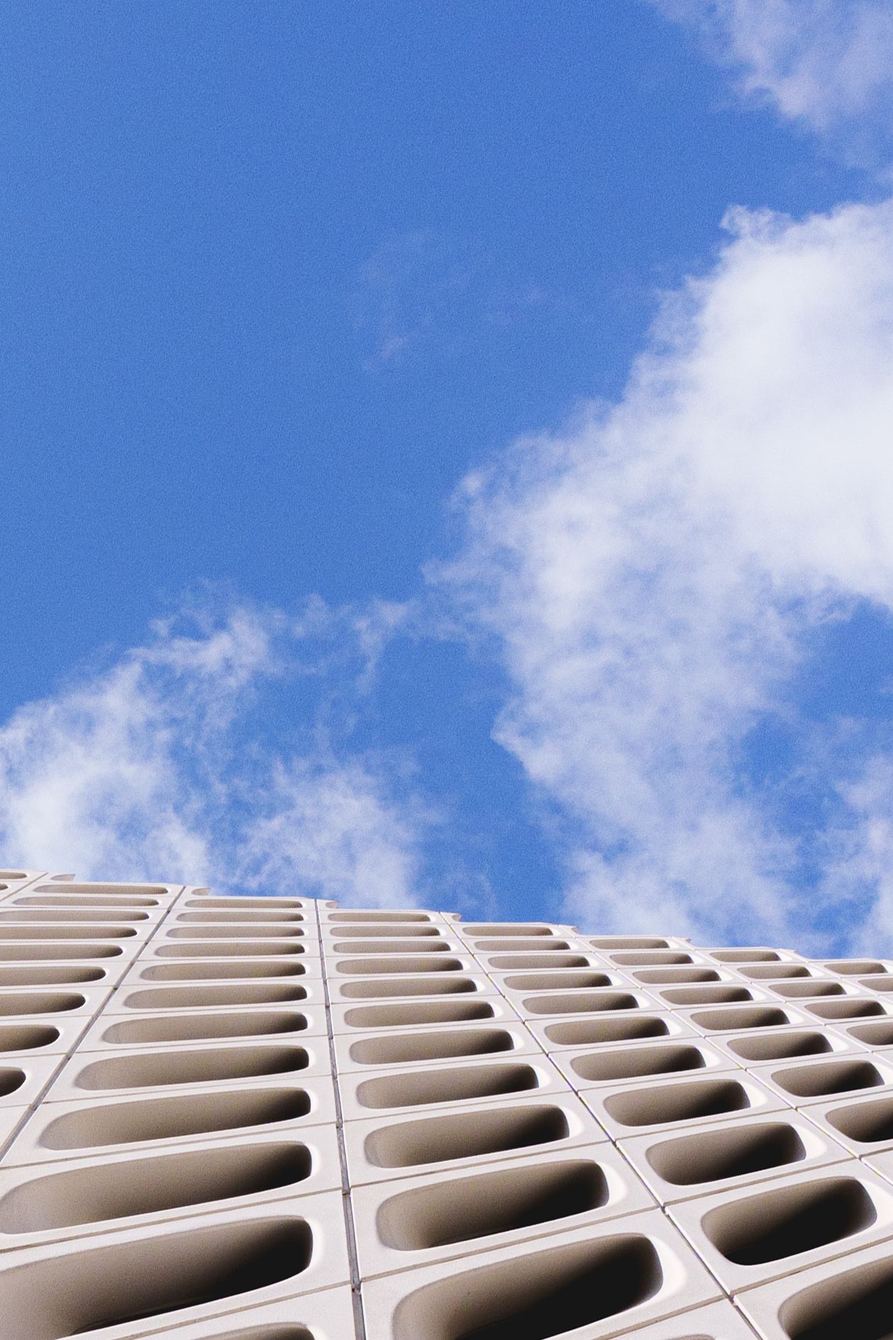 Low-angle view of a modern concrete building facade with geometric oval cutouts against a bright blue cloudy sky.