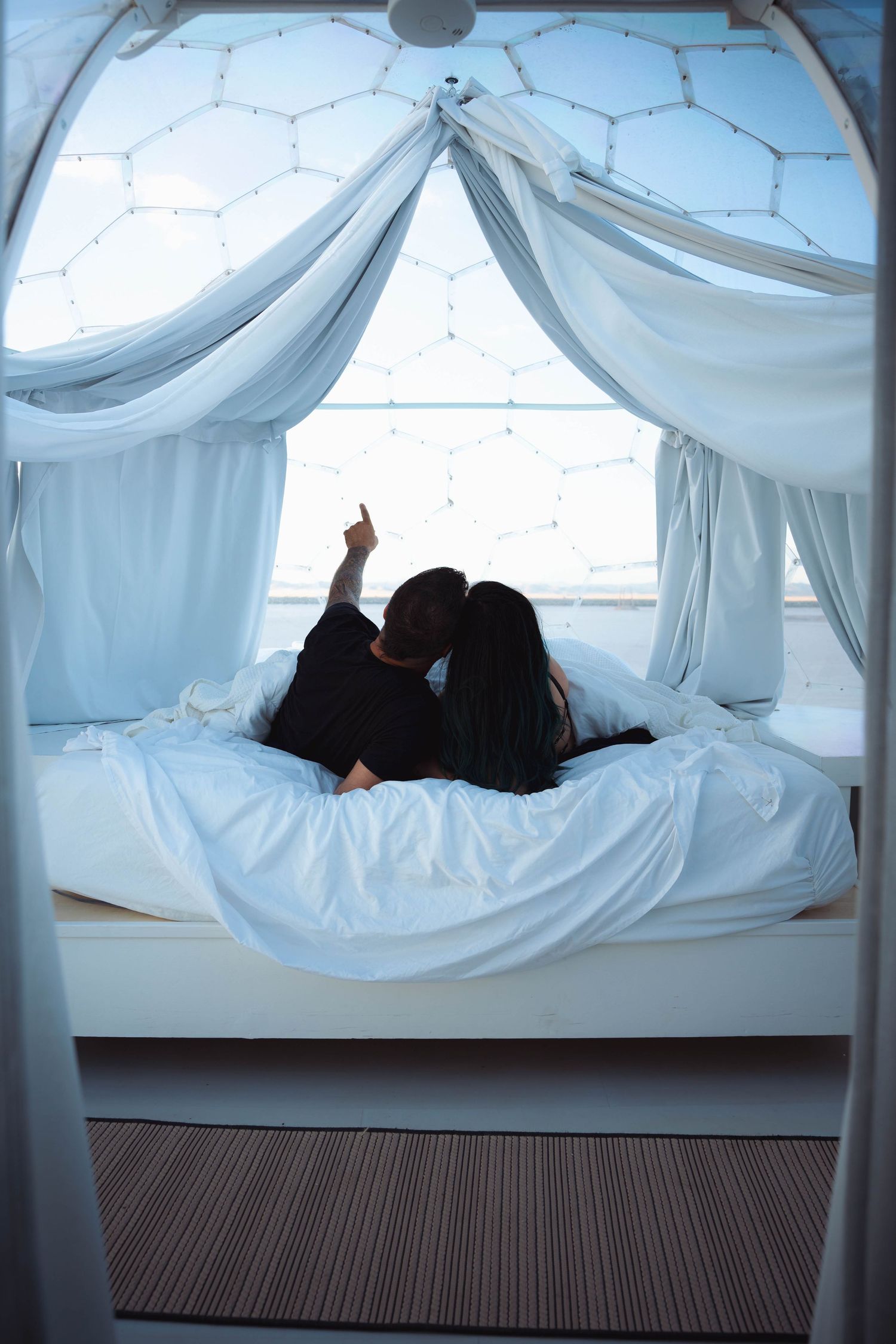 Couple lying on white bed in a dome glamping tent pointing at the ceiling.