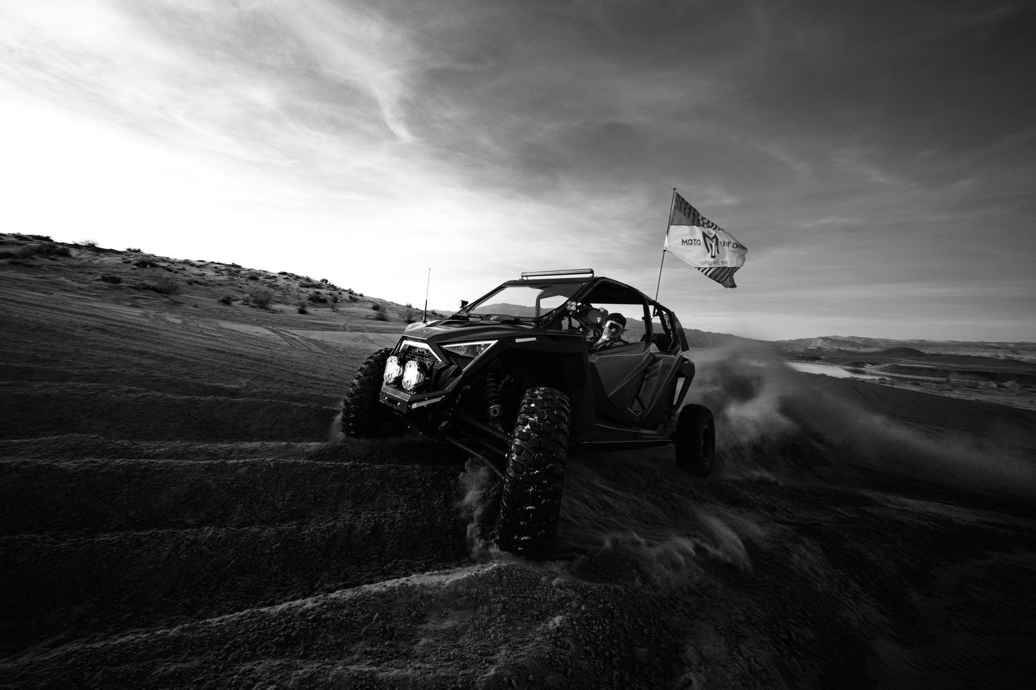 Black and white action shot of a Can-Am UTV racing across sandy desert dunes with a flag.