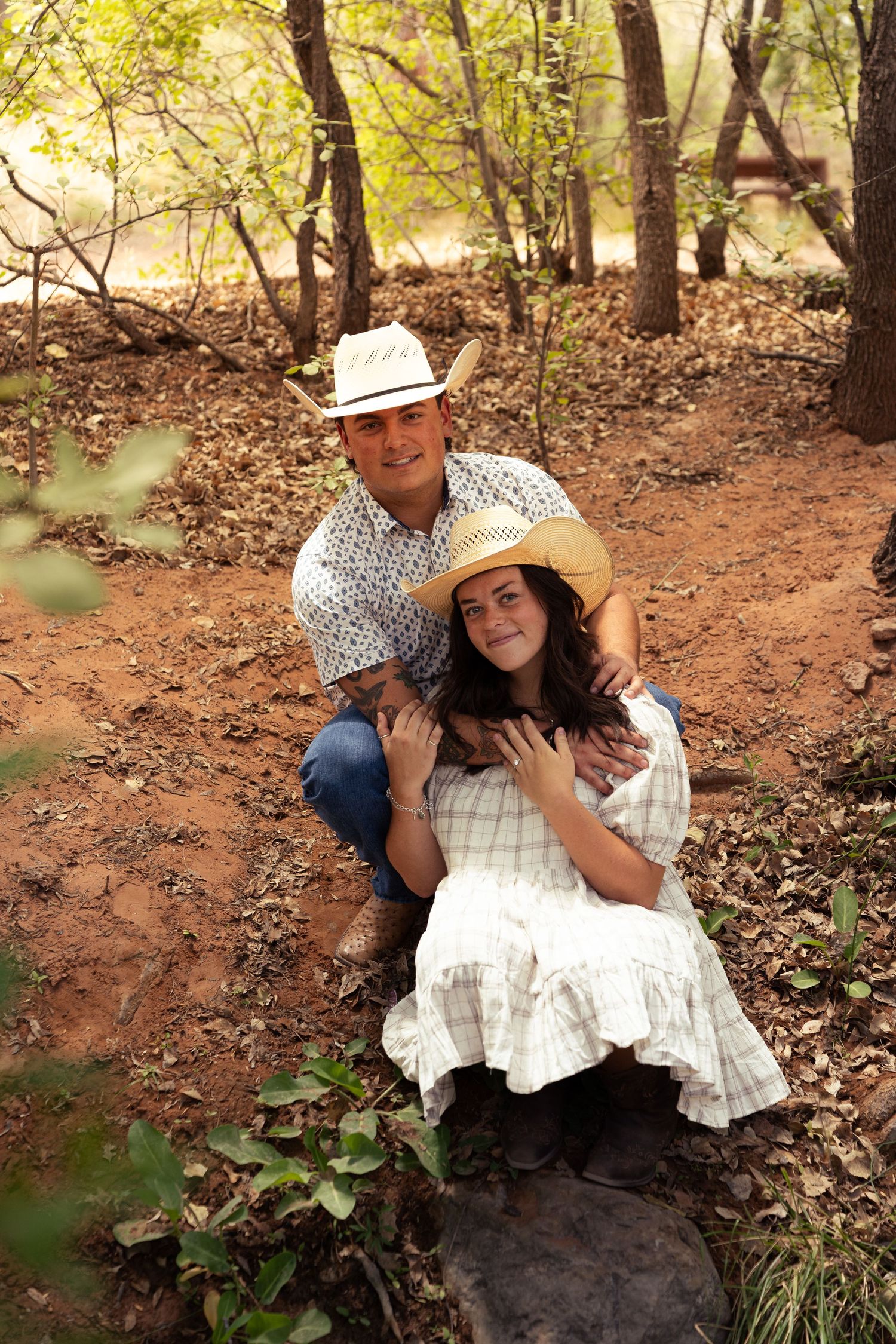 Couple in cowboy hats posing in a wooded forest setting with red dirt ground.