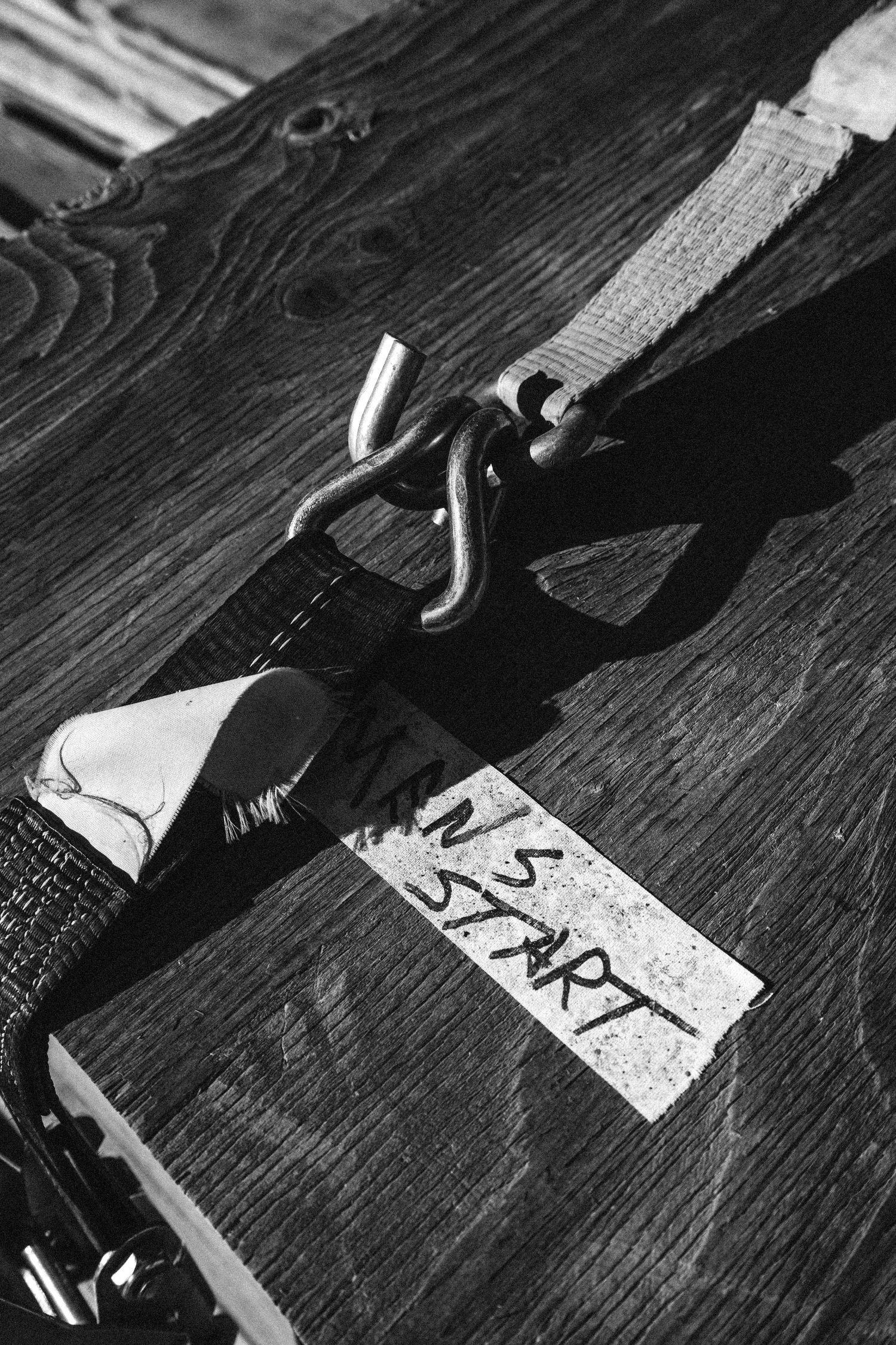 Close-up black and white photo of a ratchet strap labeled N S START secured to weathered wooden planks.