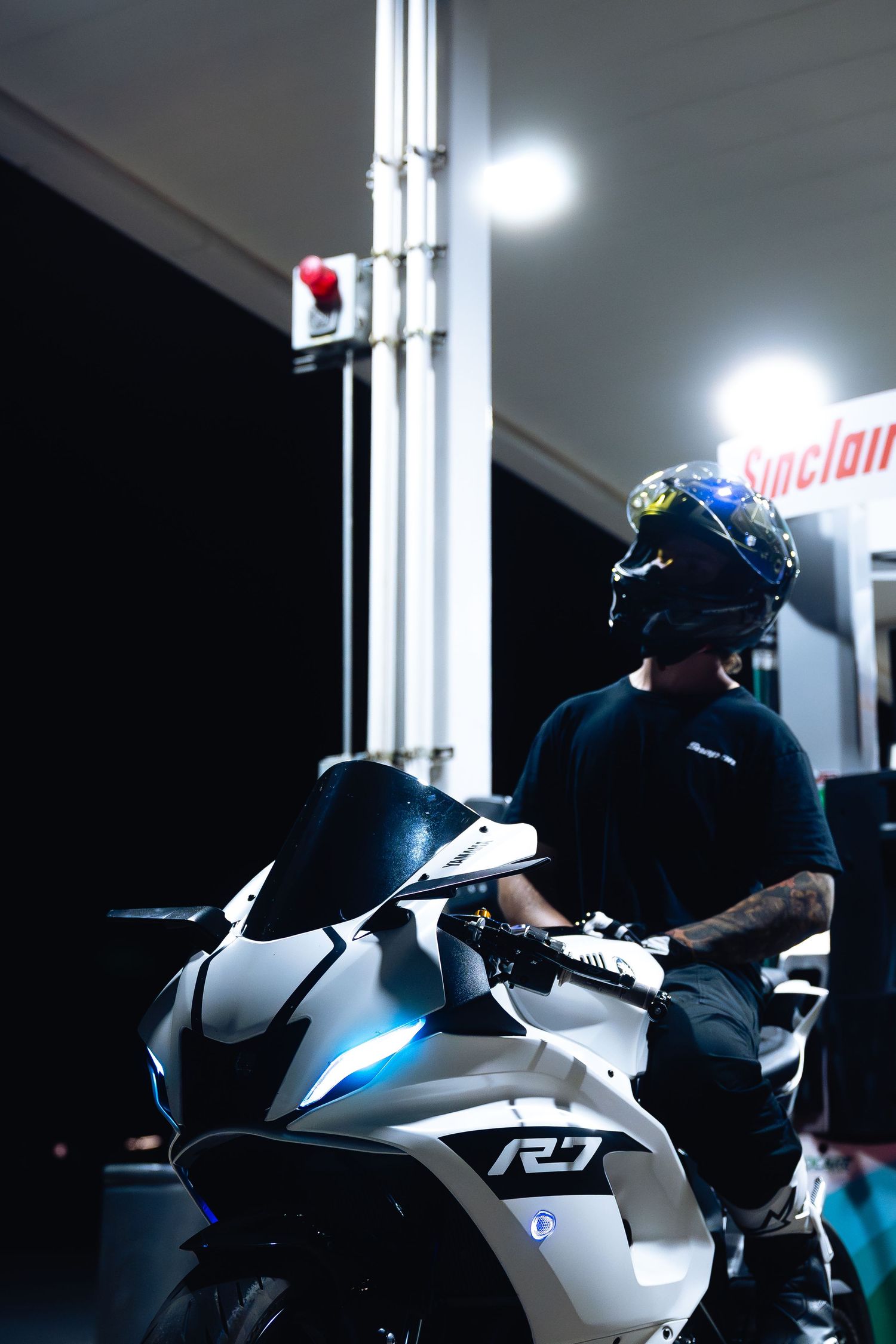 Tattooed motorcyclist in decorative helmet sits on white Yamaha R7 at Sinclair gas station.