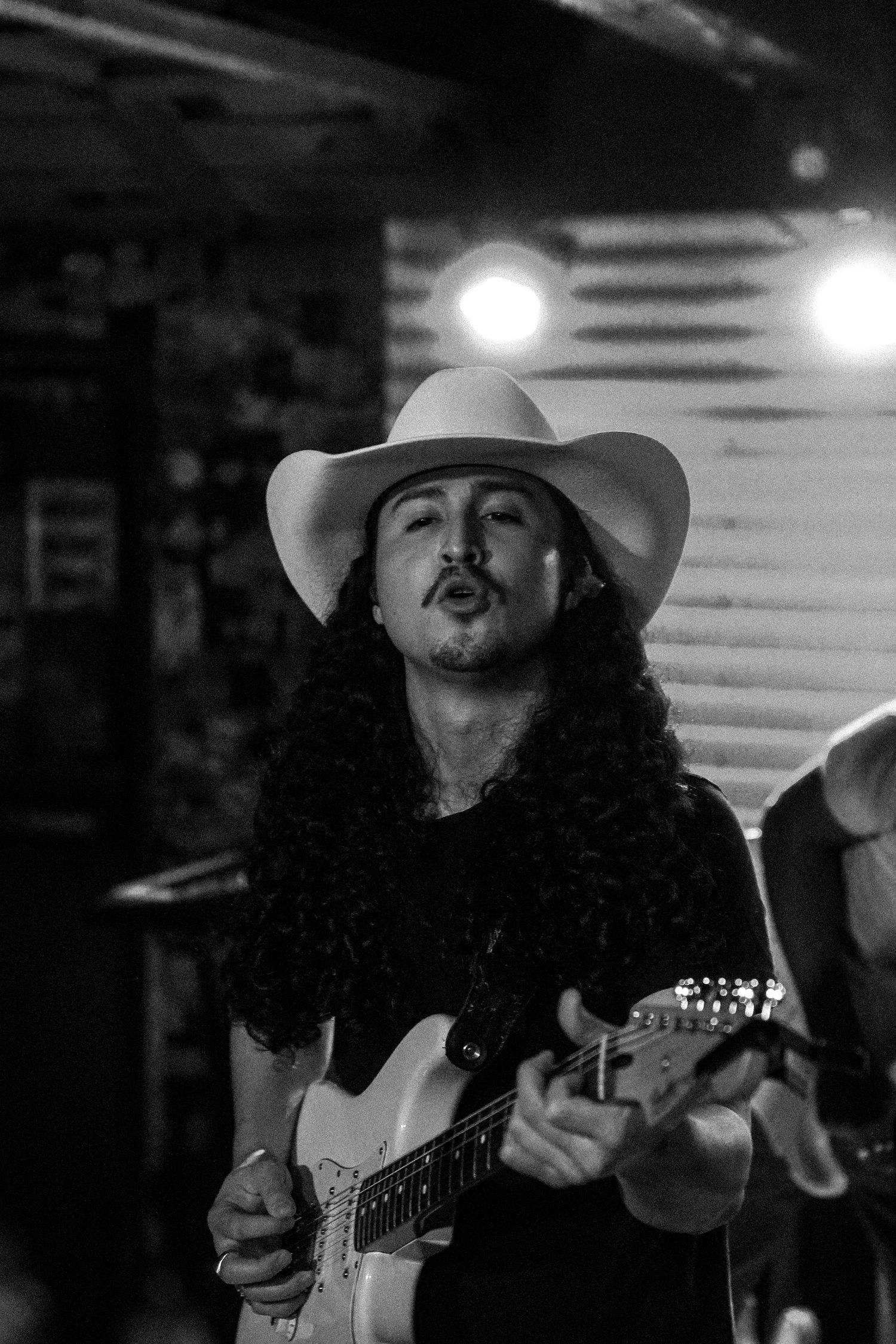 Black and white close-up of long-haired guitarist in cowboy hat singing while playing electric guitar.