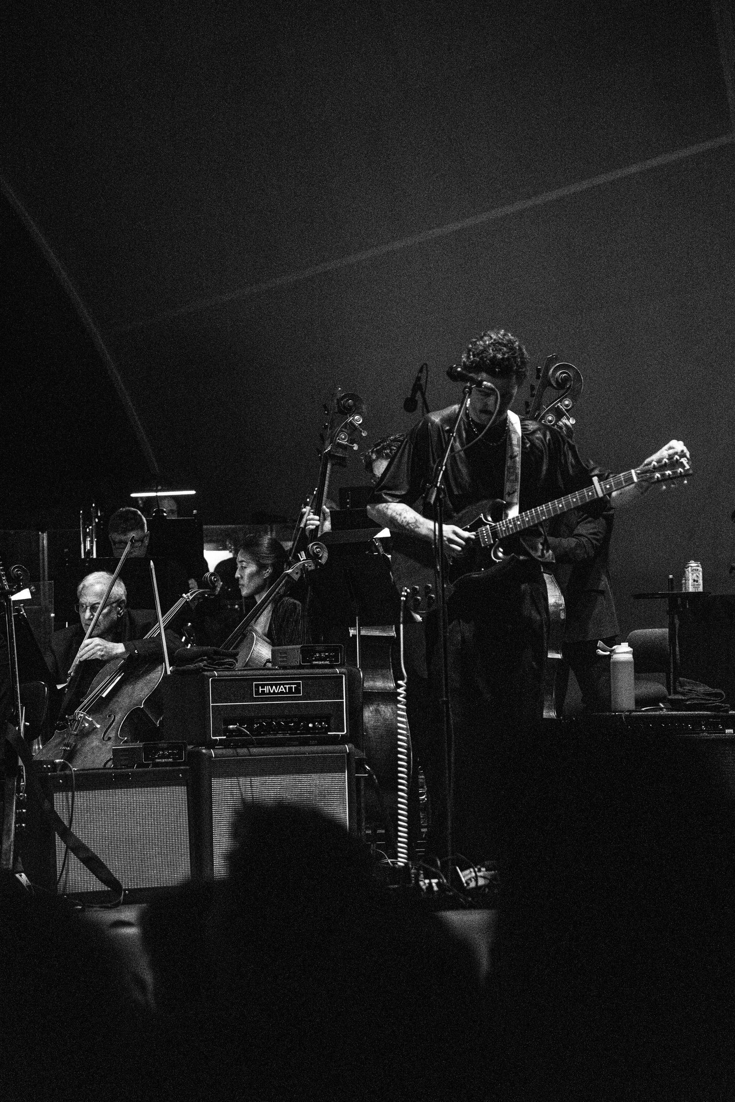 Black and white photo of a guitarist performing on stage with amplifiers and orchestra musicians visible behind them.