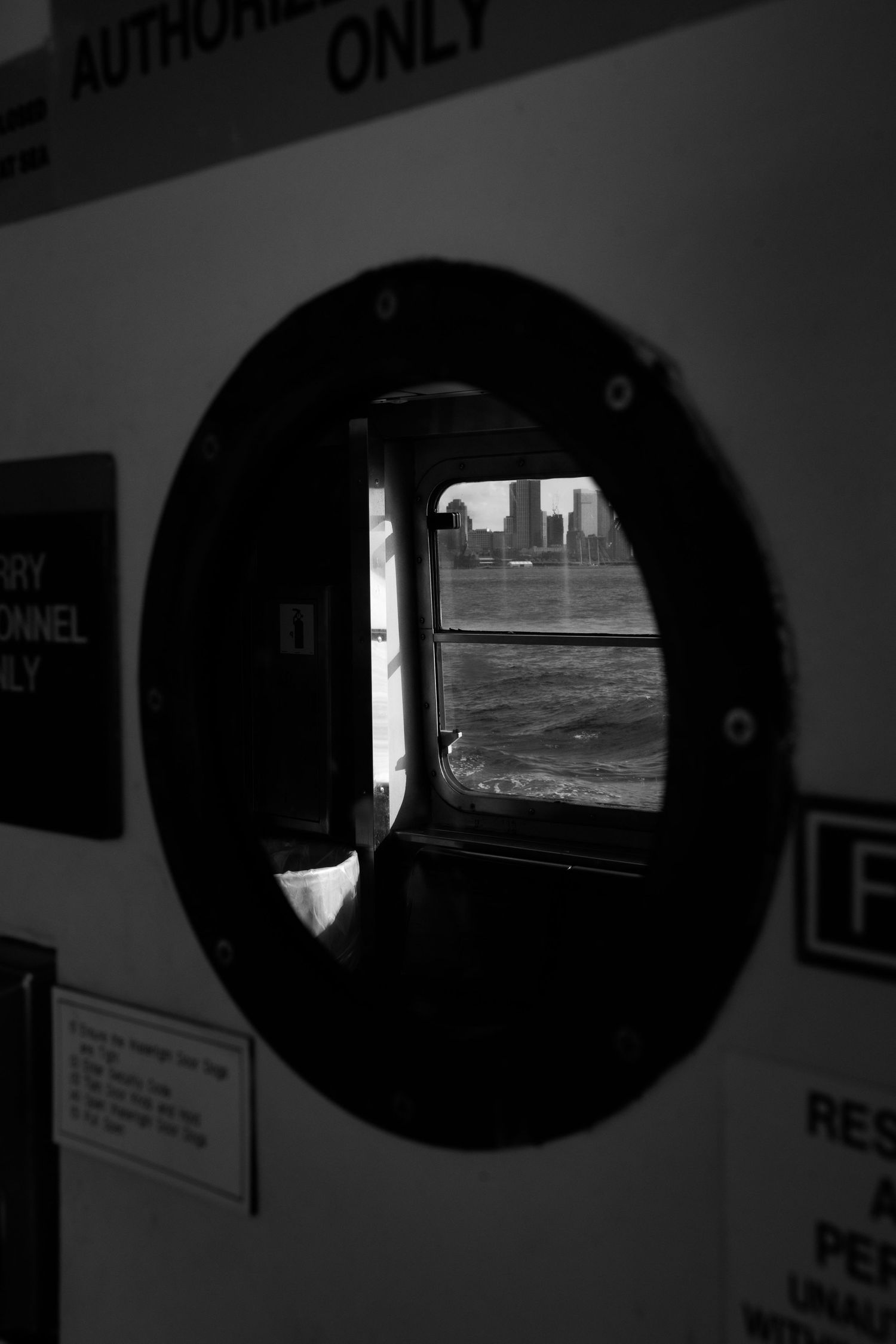 Black and white photo of a city skyline viewed through a circular porthole window on a ferry boat.